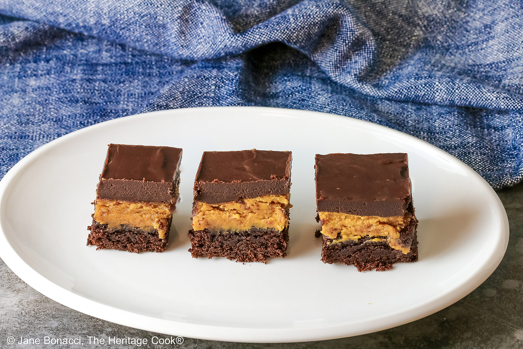 3 Layered Peanut Butter Brownies on an oval white platter with a blue cloth behind and a gray mottled background; brownie on the bottom, peanut butter layer in the middle and finished with a thick ganache chocolate layer on top ©2025 Jane Bonacci, The Heritage Cook. 