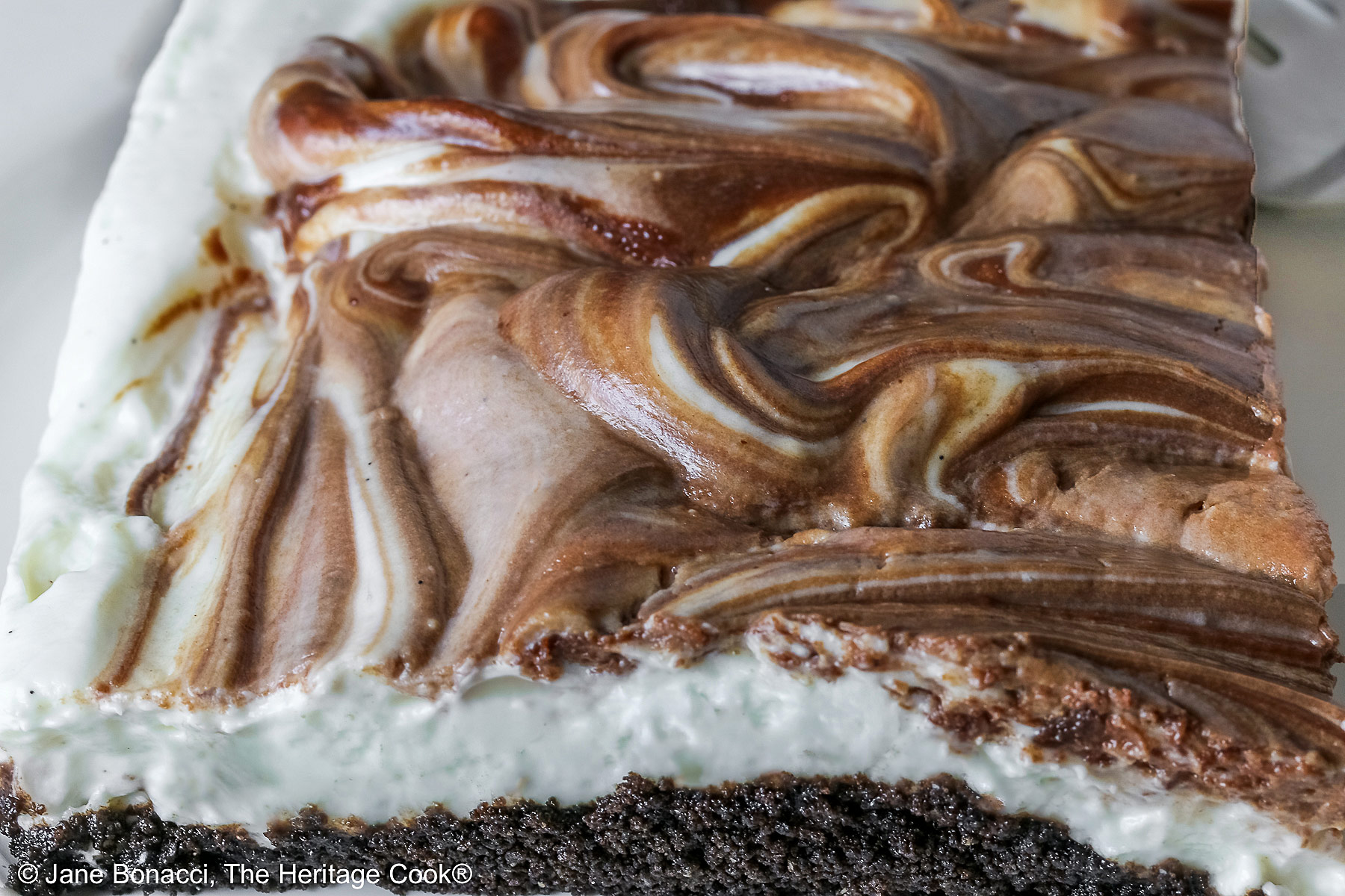 Square slice of Marbled No-Bake Cheesecake Bars on a white plate with a fork alongside, sitting on a red and white cloth on a dark gray background © 2025 Jane Bonacci, The Heritage Cook.
