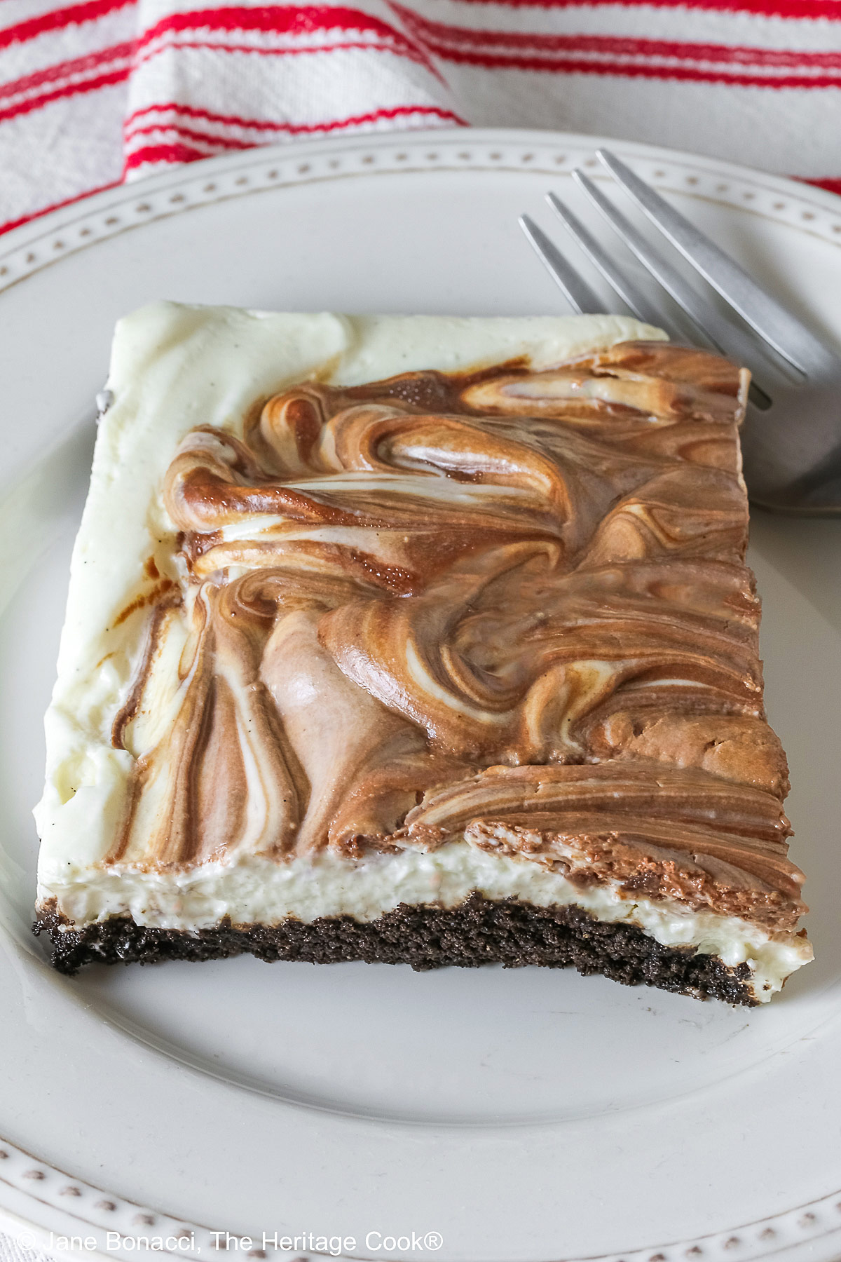 Square slice of Marbled No-Bake Cheesecake Bars on a white plate with a fork alongside, sitting on a red and white cloth on a dark gray background © 2025 Jane Bonacci, The Heritage Cook.