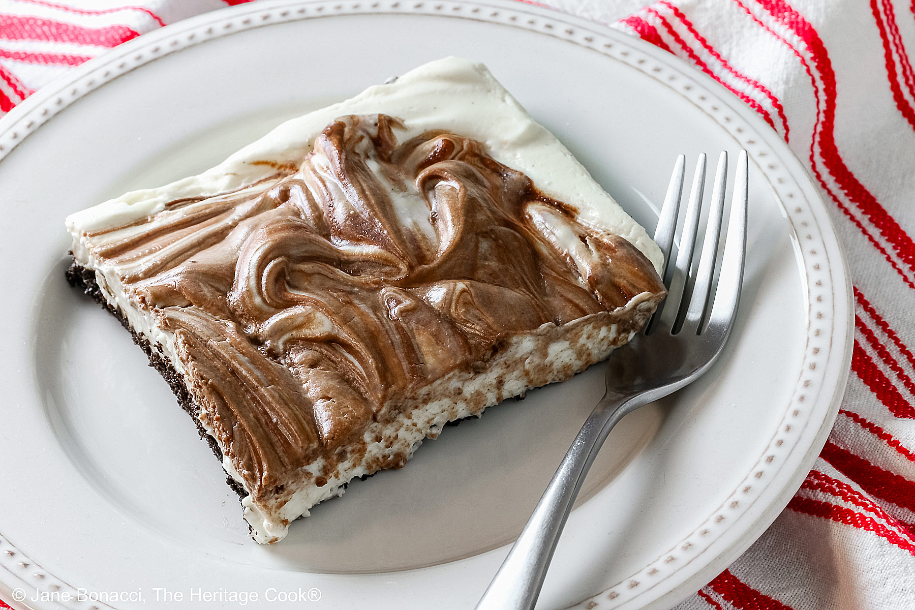 Square slice of Marbled No-Bake Cheesecake Bars on a white plate with a fork alongside, sitting on a red and white cloth on a dark gray background © 2025 Jane Bonacci, The Heritage Cook.
