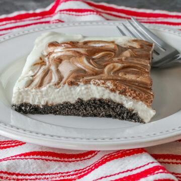Square slice of Marbled No-Bake Cheesecake Bars on a white plate with a fork alongside, sitting on a red and white cloth on a dark gray background © 2025 Jane Bonacci, The Heritage Cook.