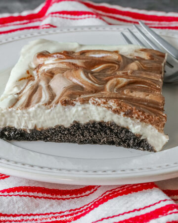 Square slice of Marbled No-Bake Cheesecake Bars on a white plate with a fork alongside, sitting on a red and white cloth on a dark gray background © 2025 Jane Bonacci, The Heritage Cook.