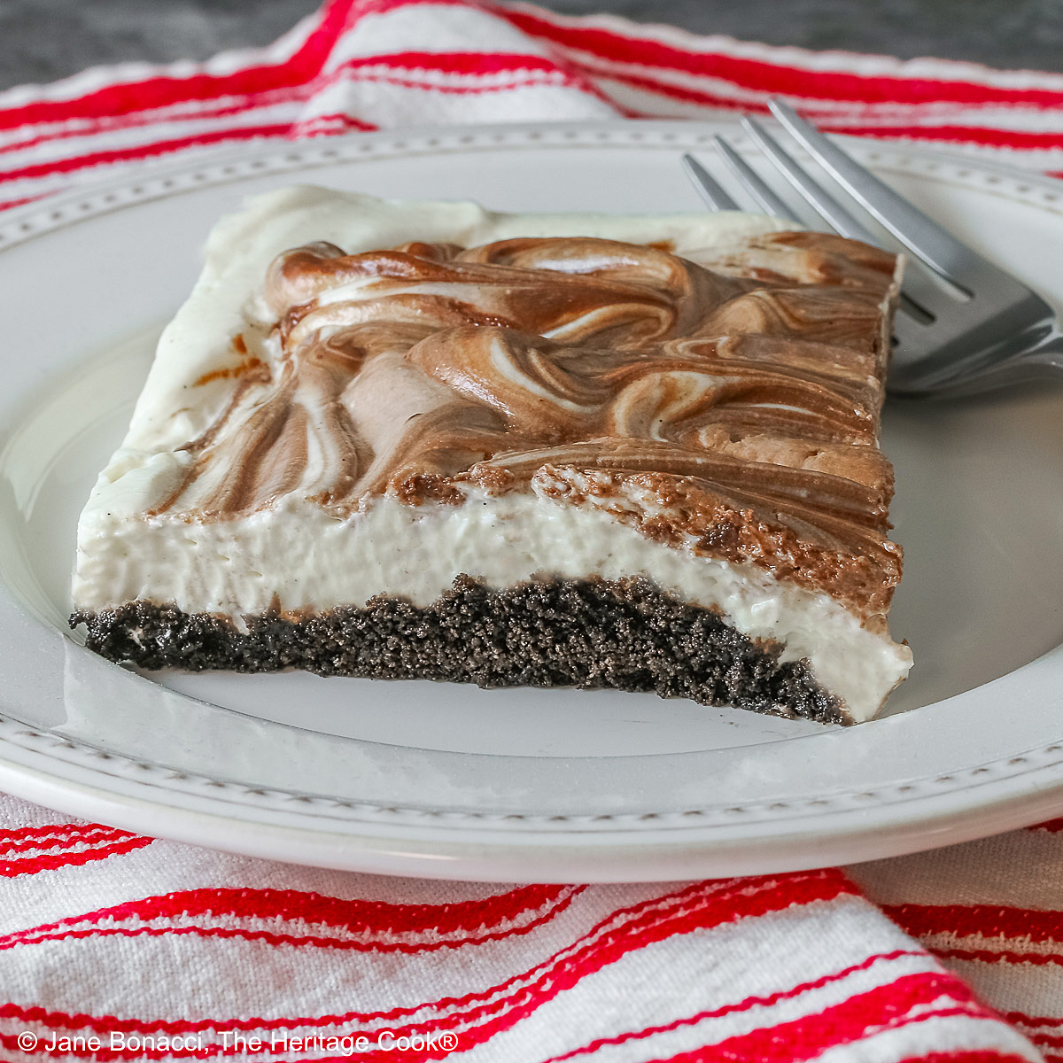Square slice of Marbled No-Bake Cheesecake Bars on a white plate with a fork alongside, sitting on a red and white cloth on a dark gray background © 2025 Jane Bonacci, The Heritage Cook.