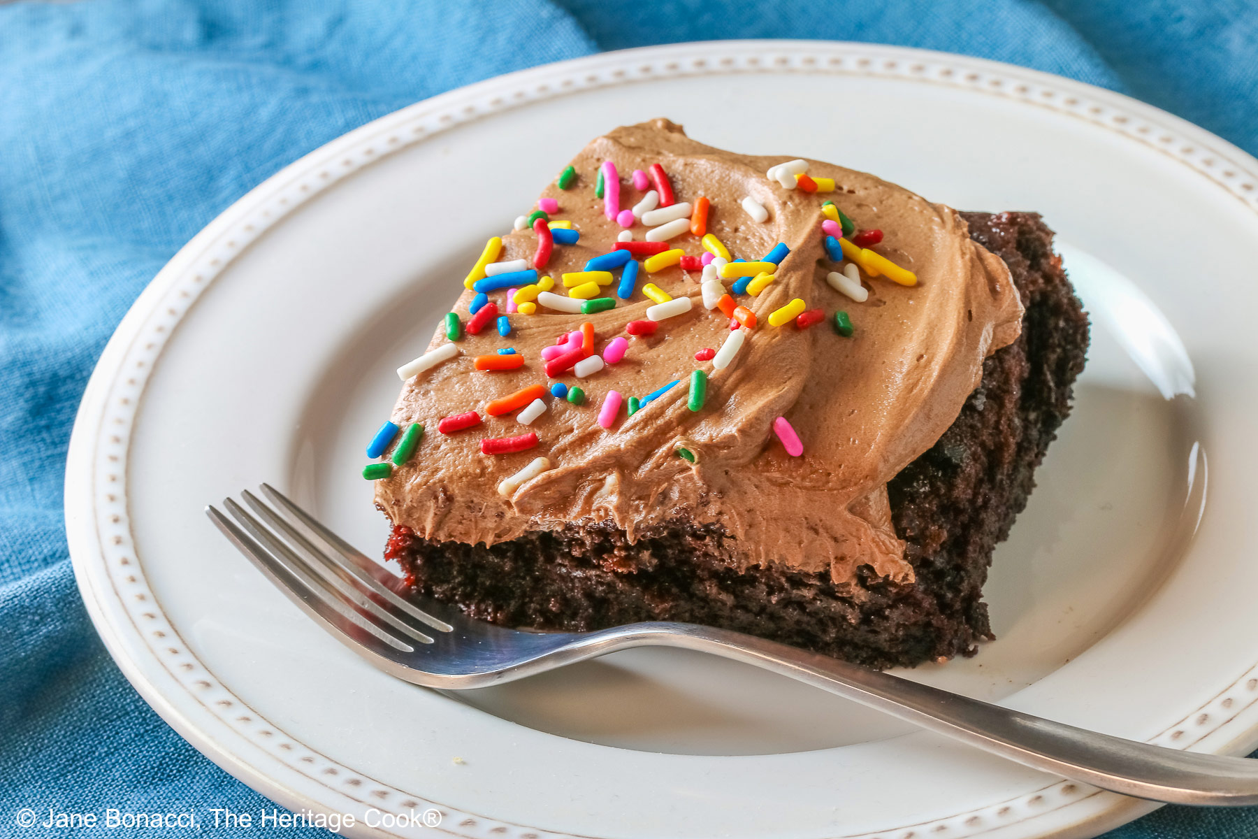 A square slice of the Easy Chocolate Sheet Cake with Chocolate Frosting on a white plate with a blue cloth beneath it and a gray background © 2025 Jane Bonacci, The Heritage Cook.