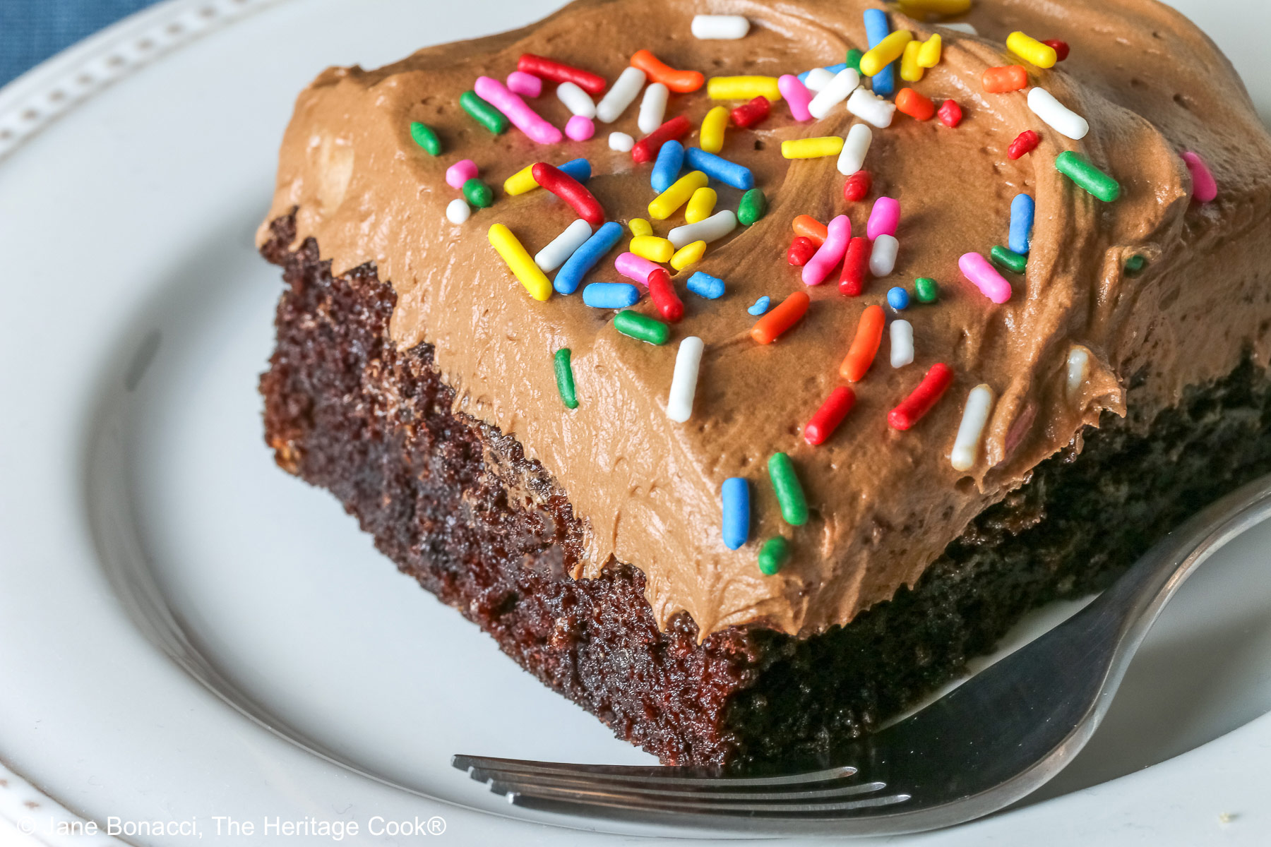 A square slice of the Easy Chocolate Sheet Cake with Chocolate Frosting on a white plate with a blue cloth beneath it and a gray background © 2025 Jane Bonacci, The Heritage Cook.