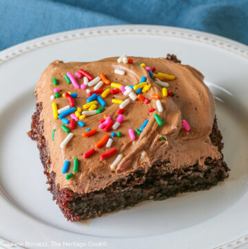 A square slice of the Easy Chocolate Sheet Cake with Chocolate Frosting on a white plate with a blue cloth beneath it and a gray background © 2025 Jane Bonacci, The Heritage Cook.