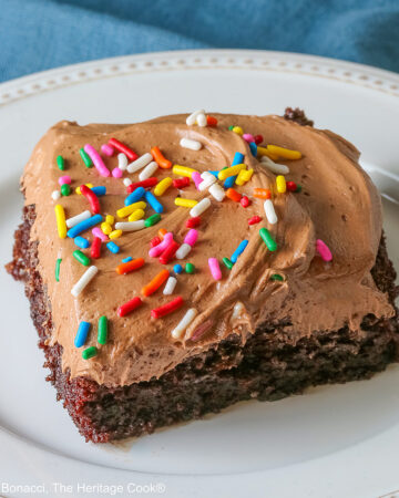 A square slice of the Easy Chocolate Sheet Cake with Chocolate Frosting on a white plate with a blue cloth beneath it and a gray background © 2025 Jane Bonacci, The Heritage Cook.