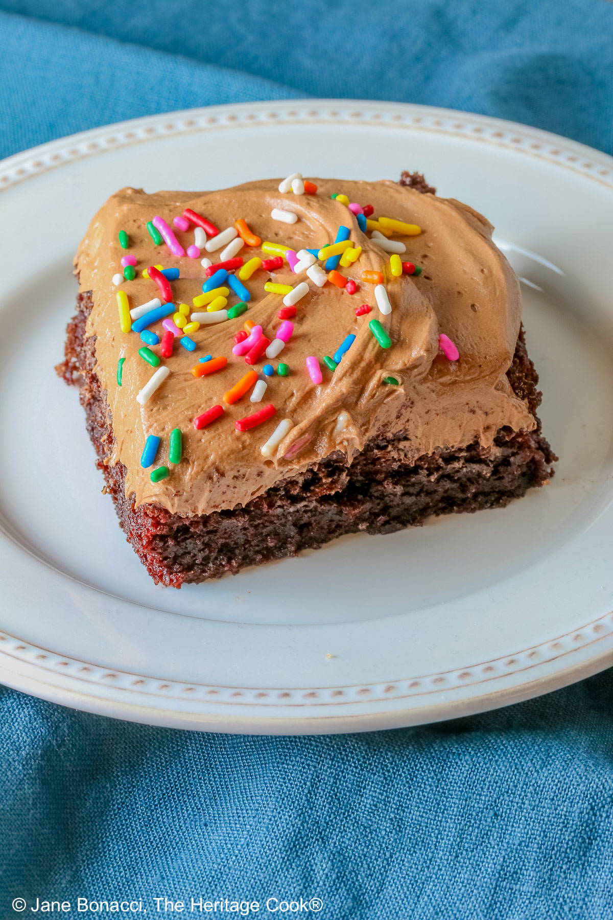A square slice of the Easy Chocolate Sheet Cake with Chocolate Frosting on a white plate with a blue cloth beneath it and a gray background © 2025 Jane Bonacci, The Heritage Cook.