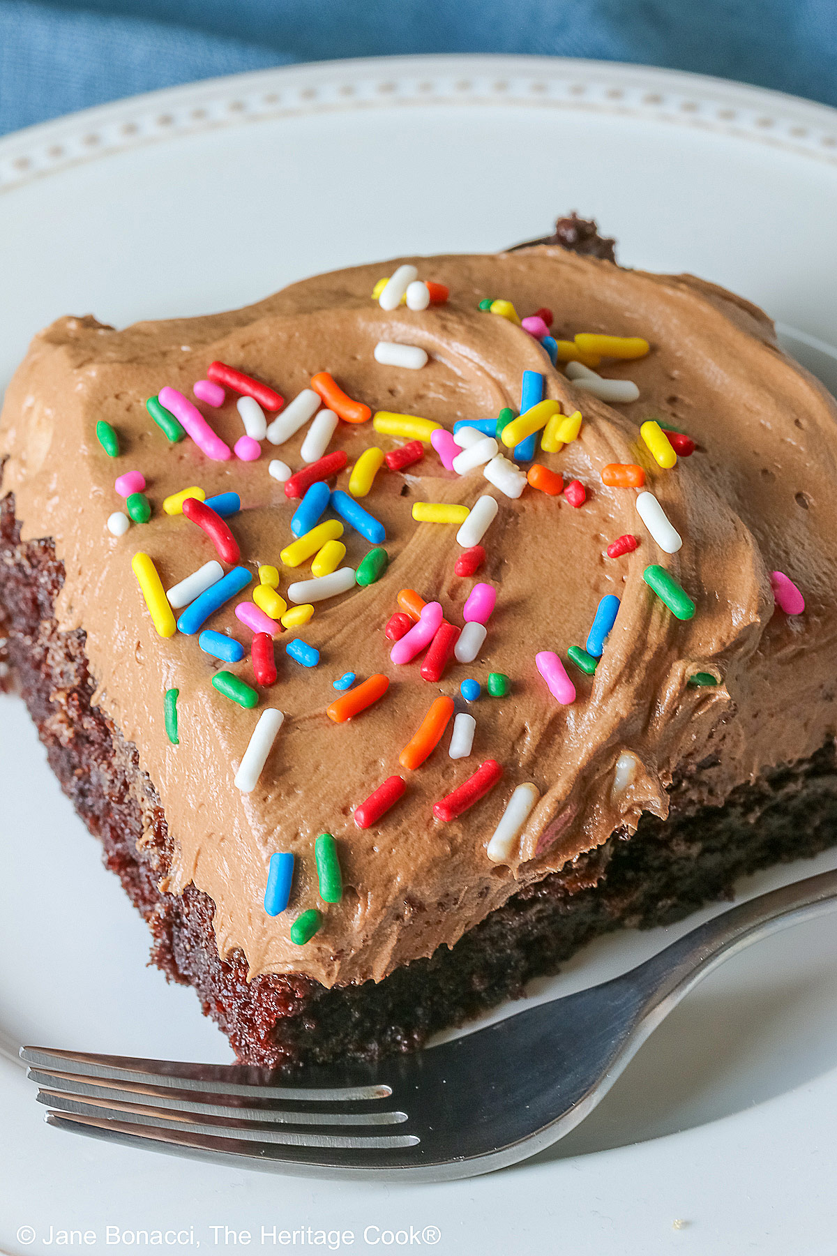 A square slice of the Easy Chocolate Sheet Cake with Chocolate Frosting on a white plate with a blue cloth beneath it and a gray background © 2025 Jane Bonacci, The Heritage Cook.