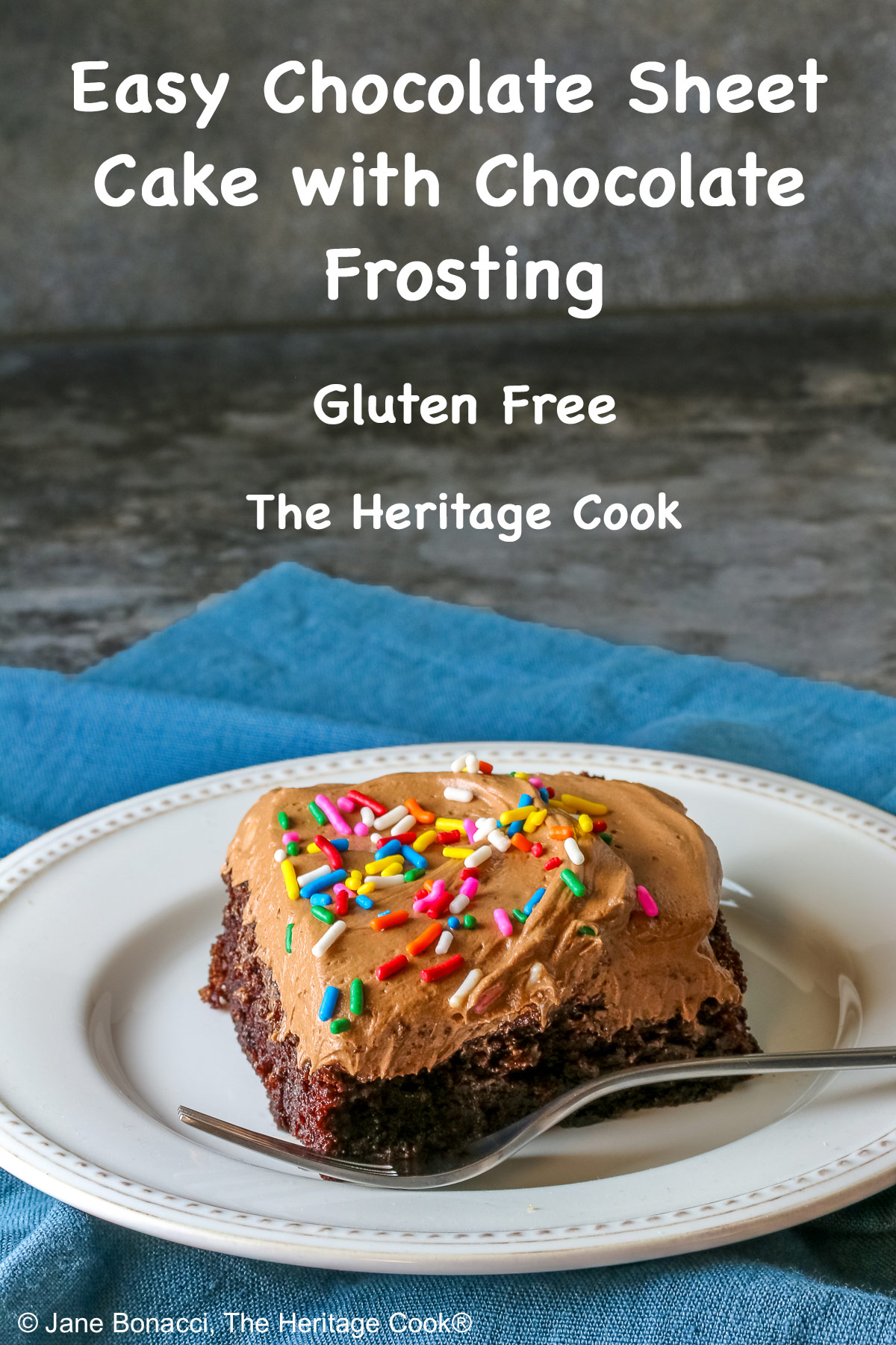 A square slice of the Easy Chocolate Sheet Cake with Chocolate Frosting on a white plate with a blue cloth beneath it and a gray background © 2025 Jane Bonacci, The Heritage Cook.