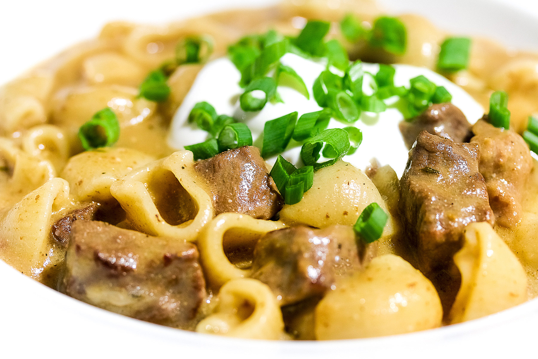 Wide rimmed white bowl filled with Easy Homemade Beef Stroganoff with pasta, beef, and sauce, topped with a dollop of sour cream and sprinkled with chopped green onions, sitting on a black background © 2025 Jane Bonacci, The Heritage Cook.
