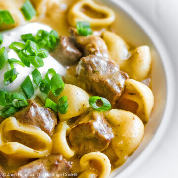 Wide rimmed white bowl filled with Easy Homemade Beef Stroganoff with pasta, beef, and sauce, topped with a dollop of sour cream and sprinkled with chopped green onions, sitting on a black background © 2025 Jane Bonacci, The Heritage Cook.