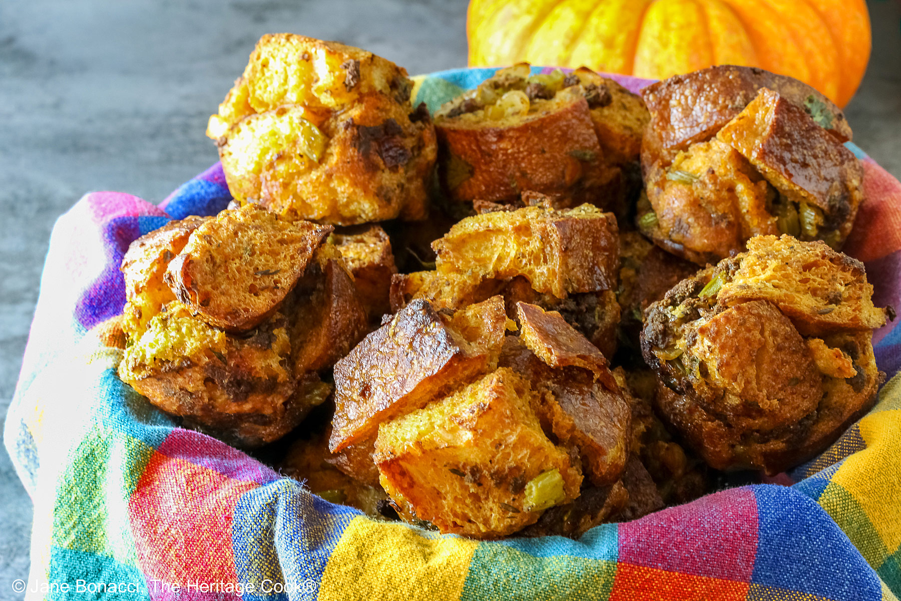 Craggy golden holiday stuffing muffins with chunks of bread and seasonings baked in a muffin tin to create round treats, tucked into a basket lined with a brightly colored cloth © 2025 Jane Bonacci, The Heritage Cook. 