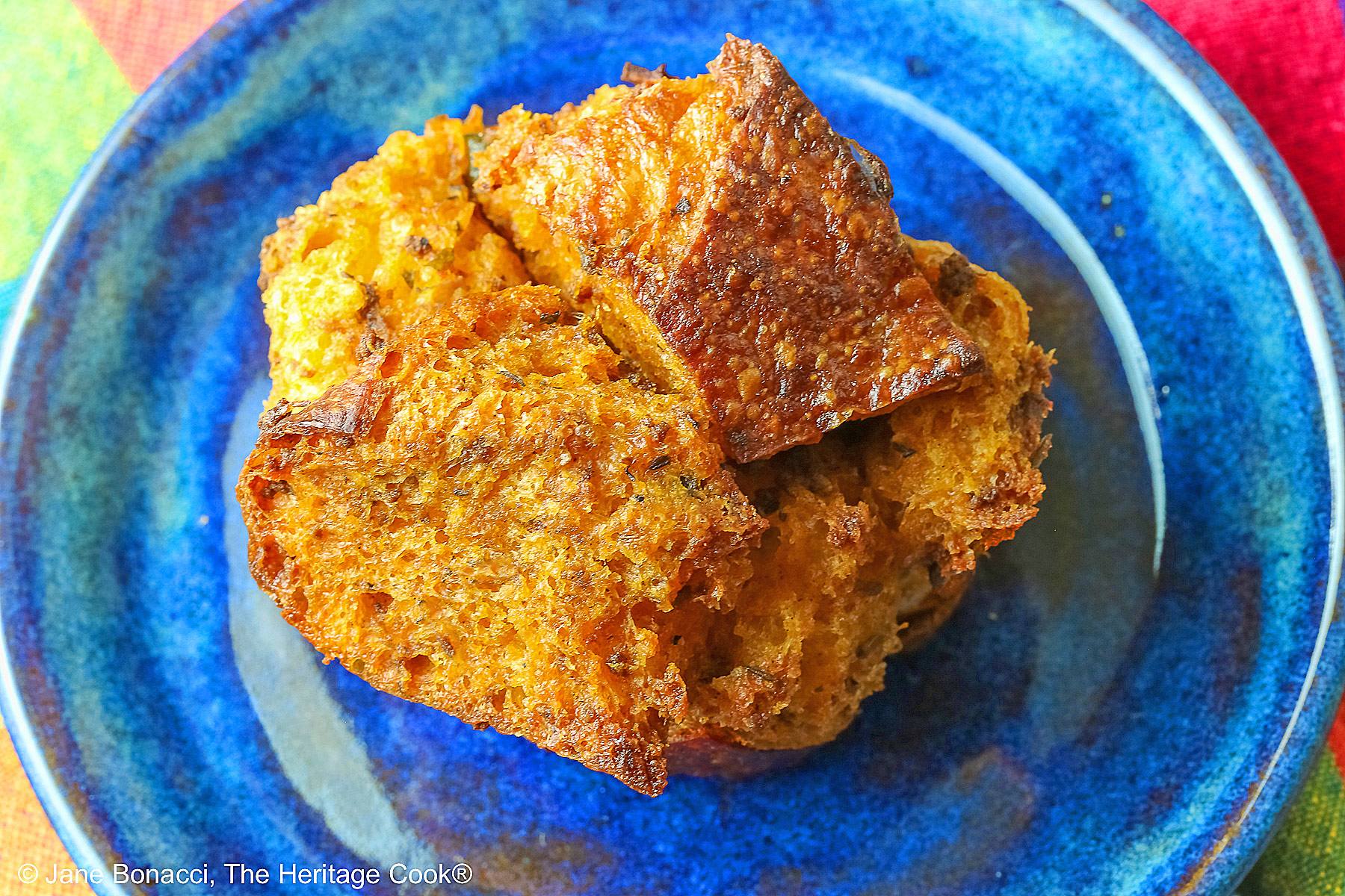 Craggy golden holiday stuffing muffins with chunks of bread and seasonings baked in a muffin tin to create round treats, tucked into a basket lined with a brightly colored cloth © 2025 Jane Bonacci, The Heritage Cook. 
