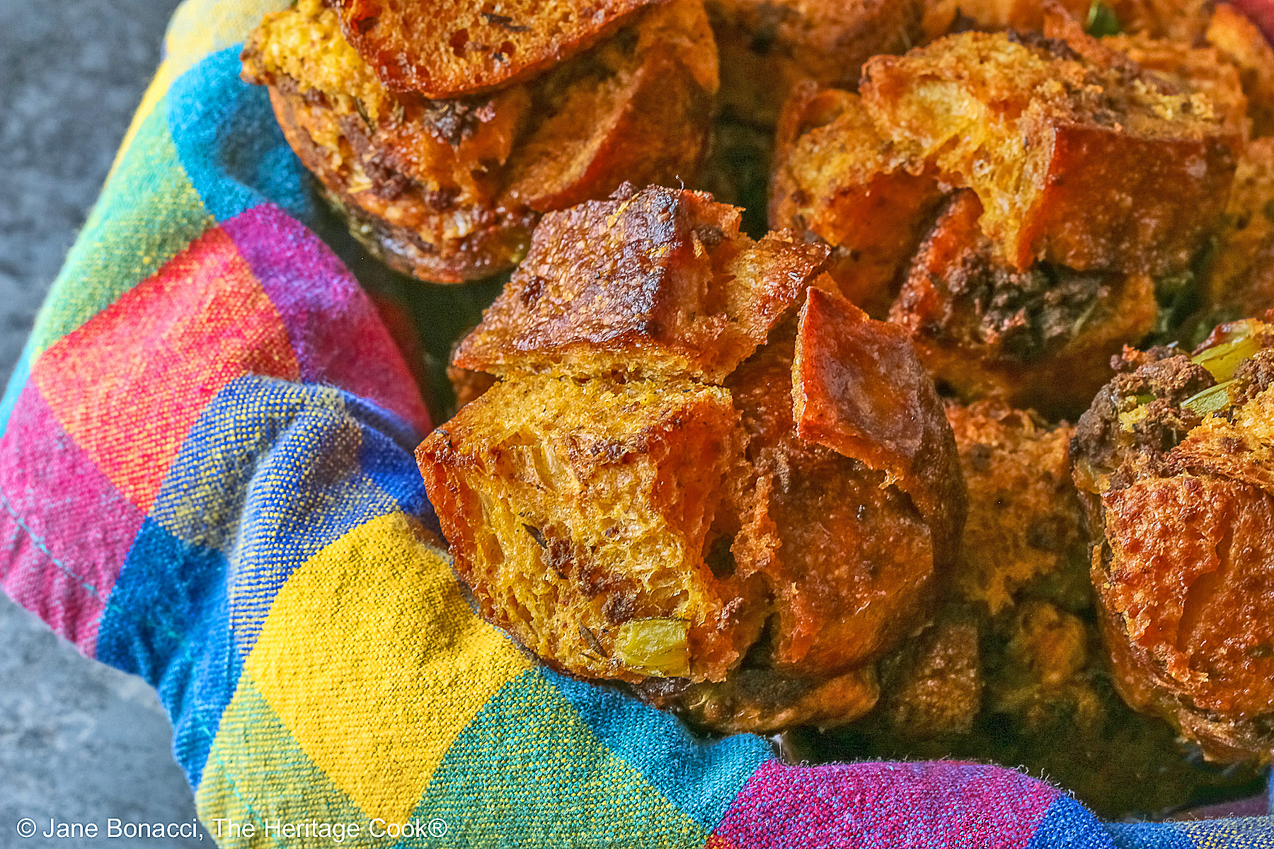 Craggy golden holiday stuffing muffins with chunks of bread and seasonings baked in a muffin tin to create round treats, tucked into a basket lined with a brightly colored cloth © 2025 Jane Bonacci, The Heritage Cook. 