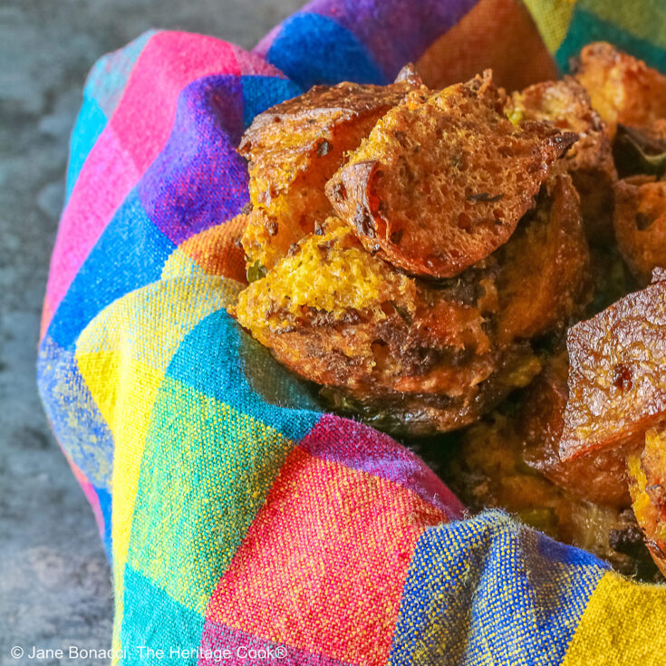 Craggy golden holiday stuffing muffins with chunks of bread and seasonings baked in a muffin tin to create round treats, tucked into a basket lined with a brightly colored cloth © 2025 Jane Bonacci, The Heritage Cook.