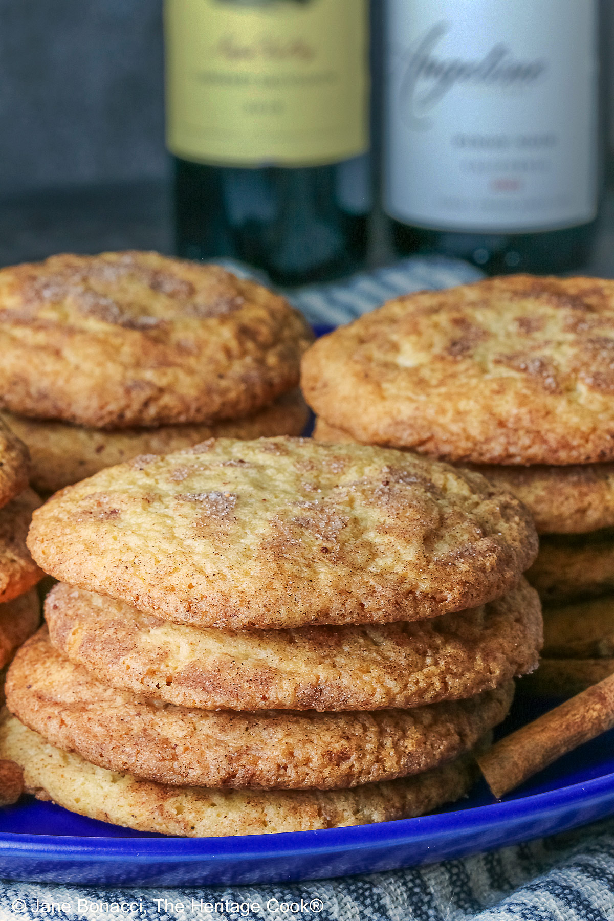 Four stacks of golden Amish White Chocolate Snickerdoodle Cookies piled on a blue plate with some cinnamon sticks crossed alongside, sitting on a pale blue striped cloth on a splattered dark gray background © 2025 Jane Bonacci, The Heritage Cook.