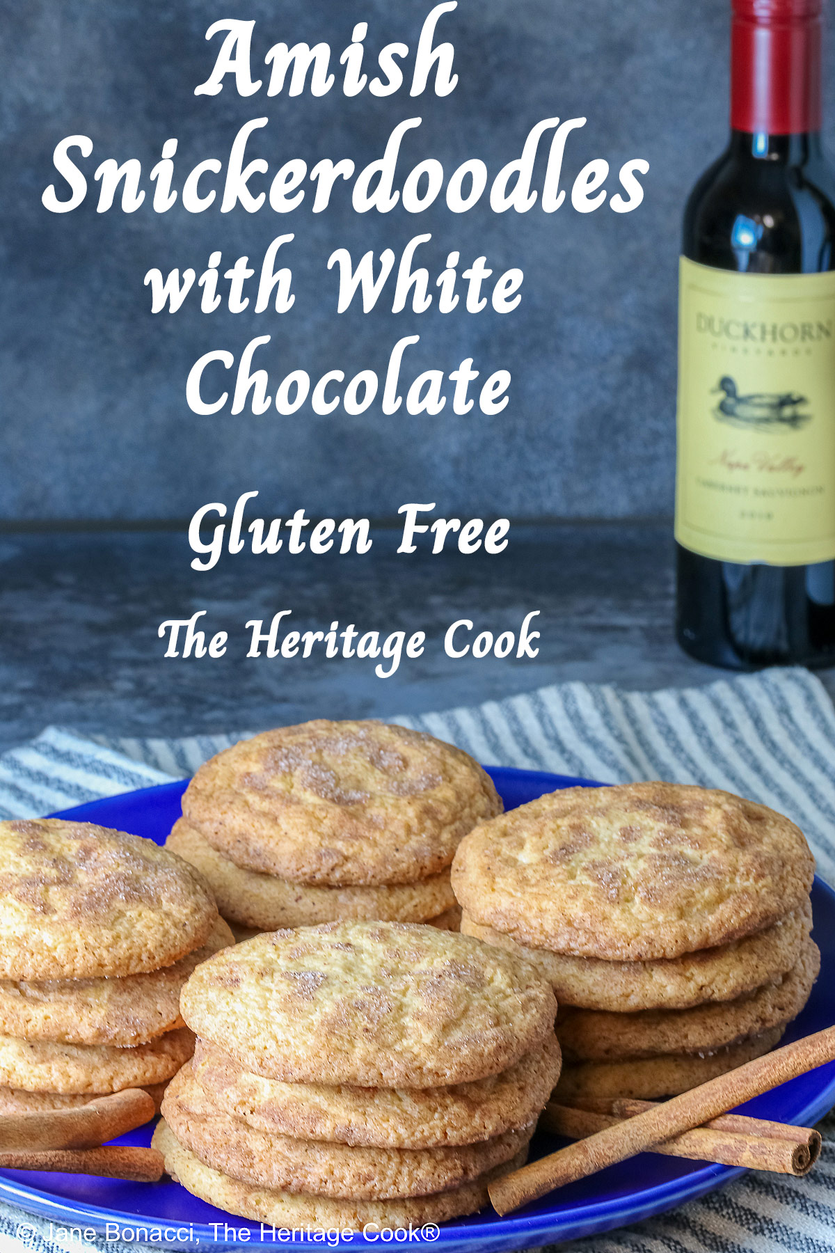 Four stacks of golden Amish White Chocolate Snickerdoodle Cookies piled on a blue plate with some cinnamon sticks crossed alongside, sitting on a pale blue striped cloth on a splattered dark gray background © 2025 Jane Bonacci, The Heritage Cook.