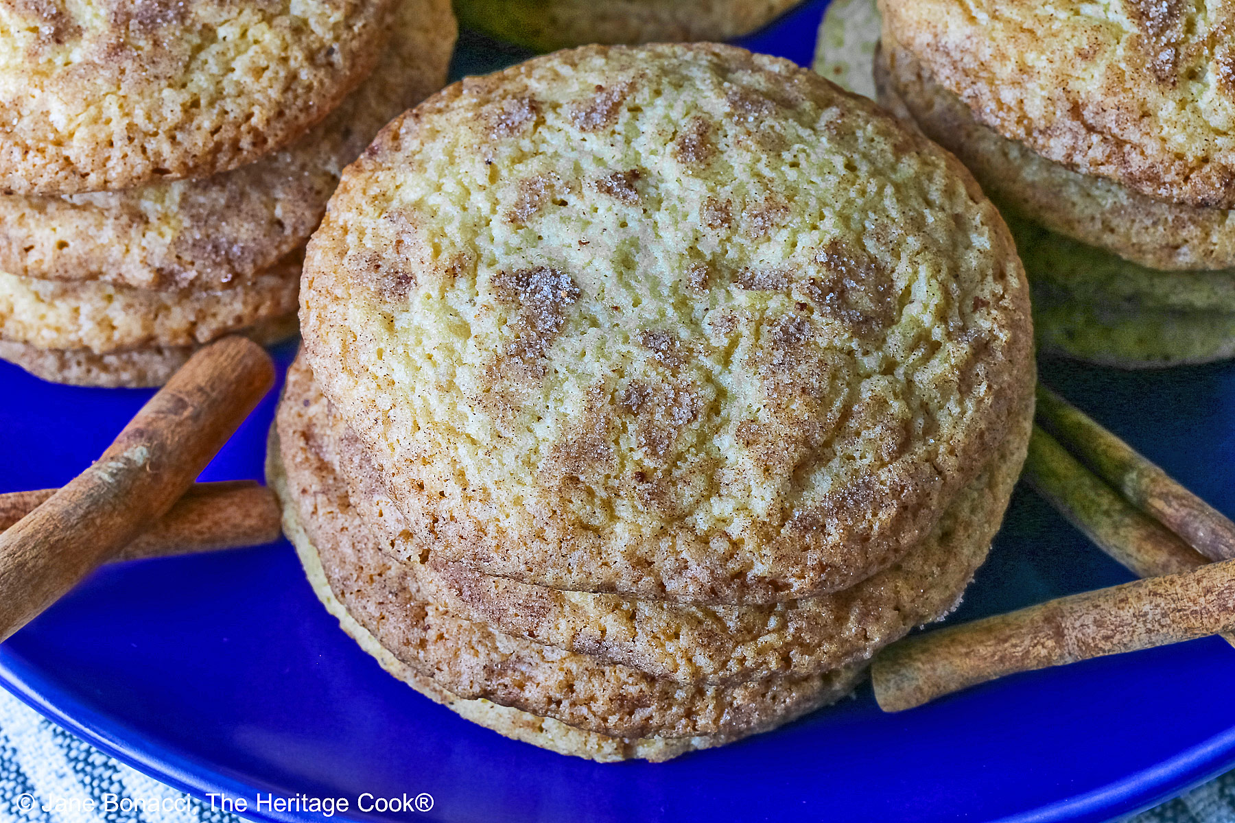 Four stacks of golden Amish White Chocolate Snickerdoodle Cookies piled on a blue plate with some cinnamon sticks crossed alongside, sitting on a pale blue striped cloth on a splattered dark gray background © 2025 Jane Bonacci, The Heritage Cook.