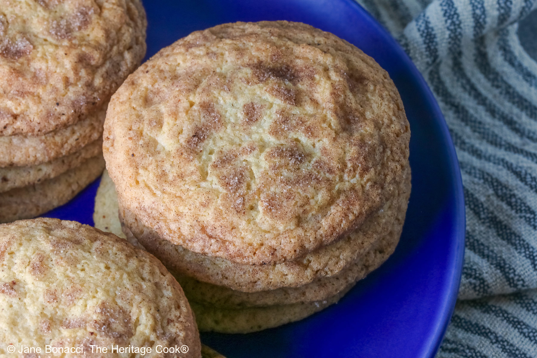 Four stacks of golden Amish White Chocolate Snickerdoodle Cookies piled on a blue plate with some cinnamon sticks crossed alongside, sitting on a pale blue striped cloth on a splattered dark gray background © 2025 Jane Bonacci, The Heritage Cook.