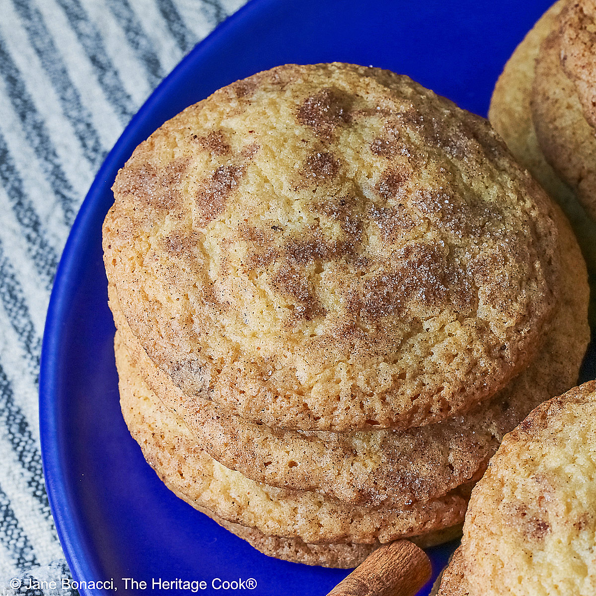 Four stacks of golden Amish White Chocolate Snickerdoodle Cookies piled on a blue plate with some cinnamon sticks crossed alongside, sitting on a pale blue striped cloth on a splattered dark gray background © 2025 Jane Bonacci, The Heritage Cook.