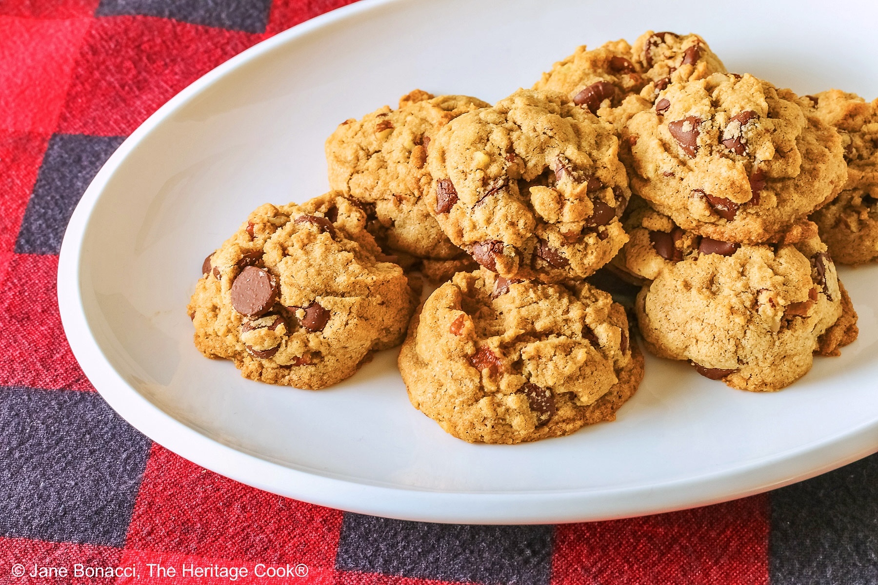 Elongated pile of Bourbon Spiked Chocolate Chip Cookies on a small white oval platter, sitting on a red and black checked buffalo plaid cloth © 2025 Jane Bonacci, The Heritage Cook.