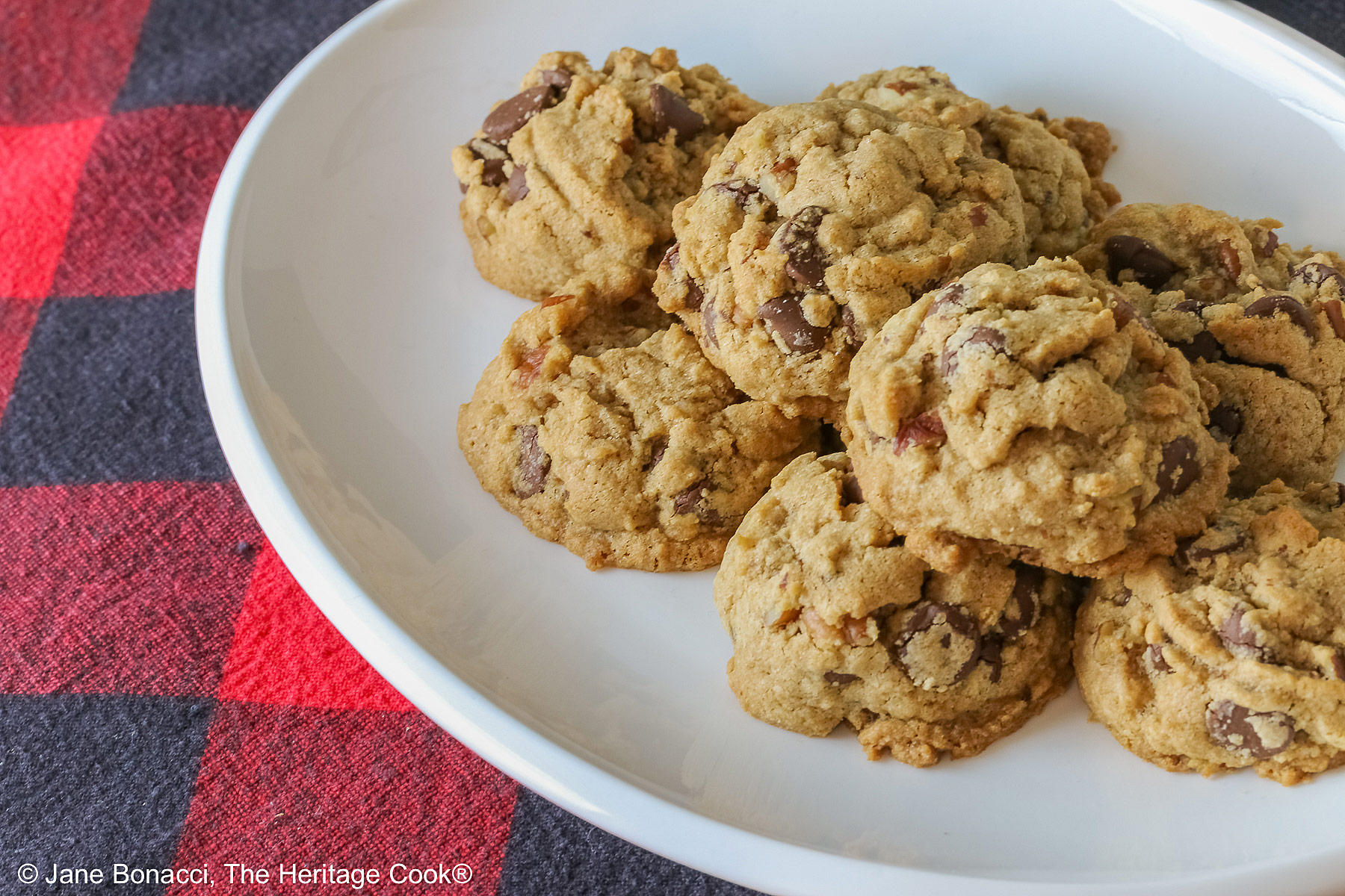 Elongated pile of Bourbon Spiked Chocolate Chip Cookies on a small white oval platter, sitting on a red and black checked buffalo plaid cloth © 2025 Jane Bonacci, The Heritage Cook.