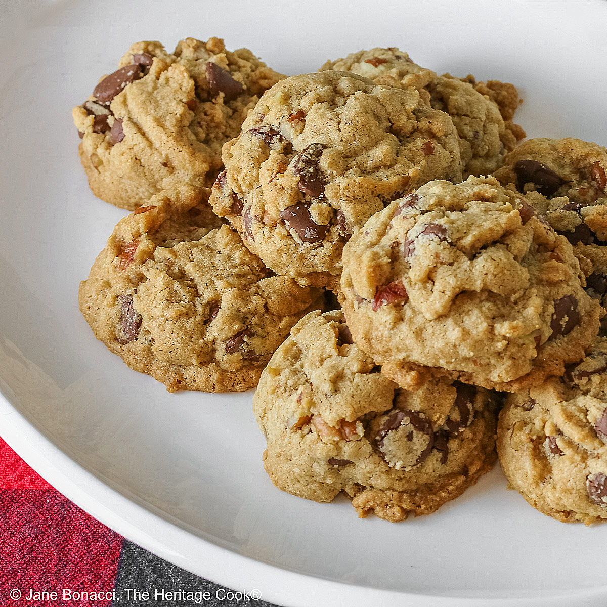 Elongated pile of Bourbon Spiked Chocolate Chip Cookies on a small white oval platter, sitting on a red and black checked buffalo plaid cloth © 2025 Jane Bonacci, The Heritage Cook.