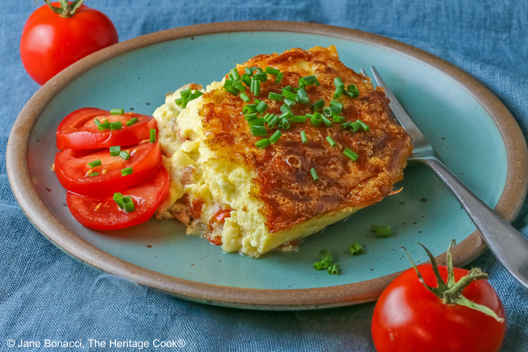 Aqua plate with a square of the crustless quiche with chopped chives on top and 3 tomato slices beside the quiche plus two whole tomatoes next to the plate, sitting on a blue cloth © 2025 Jane Bonacci, The Heritage Cook.