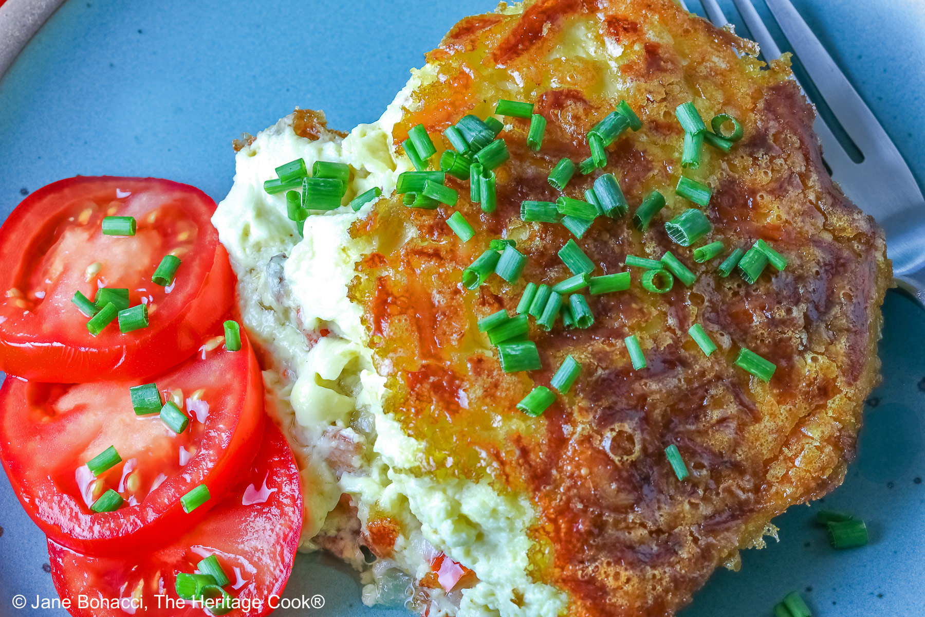 Aqua plate with a square of the crustless quiche with chopped chives on top and 3 tomato slices beside the quiche plus two whole tomatoes next to the plate, sitting on a blue cloth © 2025 Jane Bonacci, The Heritage Cook.