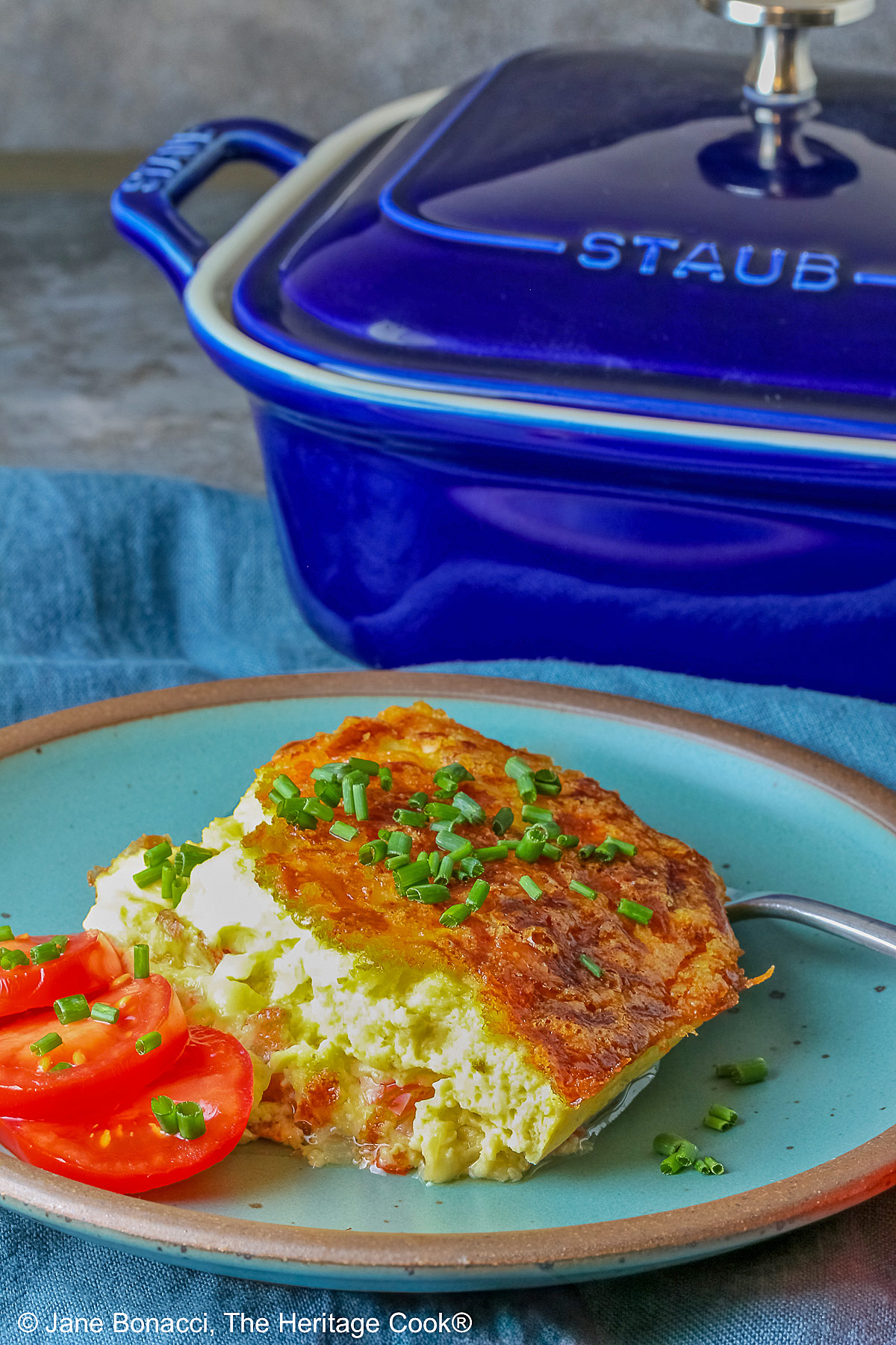 Aqua plate with a square of the crustless quiche with chopped chives on top and 3 tomato slices beside the quiche plus two whole tomatoes next to the plate, sitting on a blue cloth © 2025 Jane Bonacci, The Heritage Cook.
