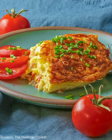 Aqua plate with a square of the crustless quiche with chopped chives on top and 3 tomato slices beside the quiche plus two whole tomatoes next to the plate, sitting on a blue cloth © 2025 Jane Bonacci, The Heritage Cook.