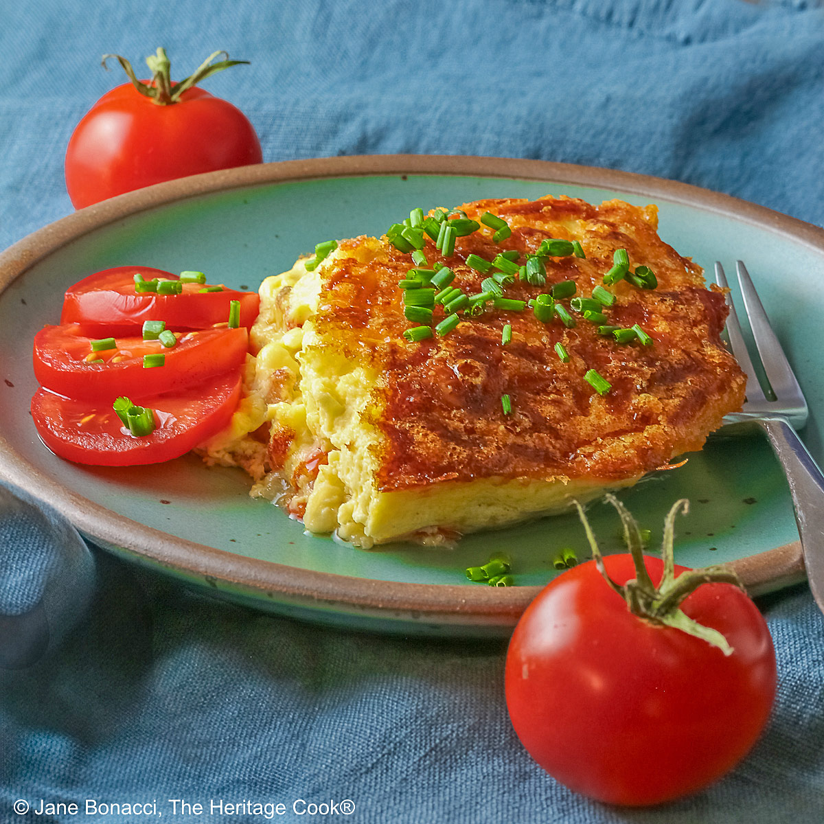 Aqua plate with a square of the crustless quiche with chopped chives on top and 3 tomato slices beside the quiche plus two whole tomatoes next to the plate, sitting on a blue cloth © 2025 Jane Bonacci, The Heritage Cook.
