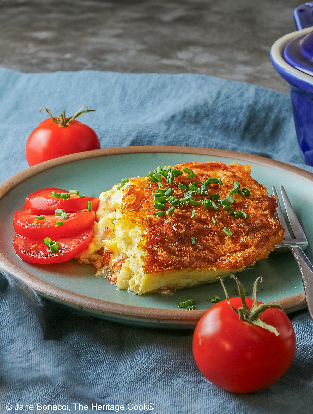 Aqua plate with a square of the crustless quiche with chopped chives on top and 3 tomato slices beside the quiche plus two whole tomatoes next to the plate, sitting on a blue cloth © 2025 Jane Bonacci, The Heritage Cook.