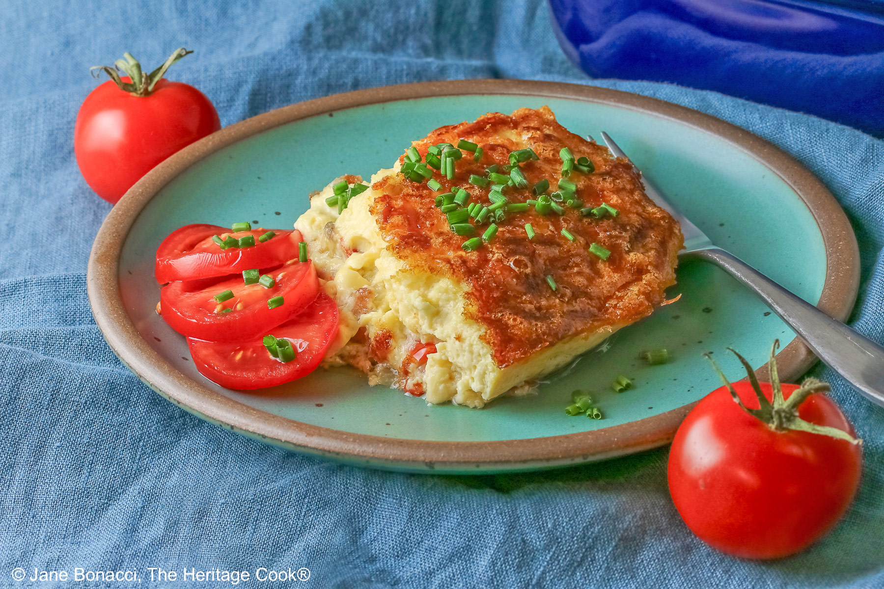 Aqua plate with a square of the crustless quiche with chopped chives on top and 3 tomato slices beside the quiche plus two whole tomatoes next to the plate, sitting on a blue cloth © 2025 Jane Bonacci, The Heritage Cook.