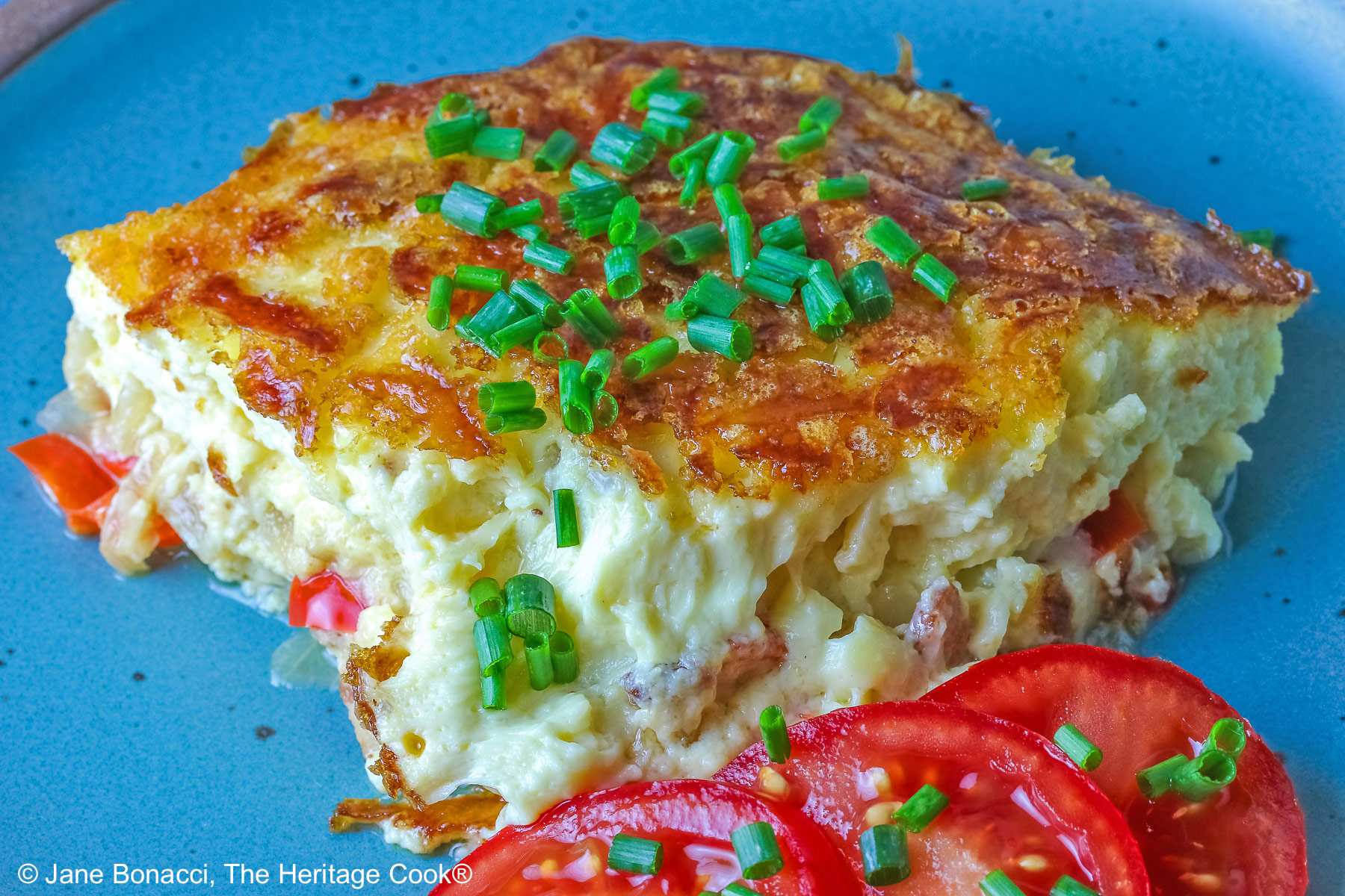 Aqua plate with a square of the crustless quiche with chopped chives on top and 3 tomato slices beside the quiche plus two whole tomatoes next to the plate, sitting on a blue cloth © 2025 Jane Bonacci, The Heritage Cook.