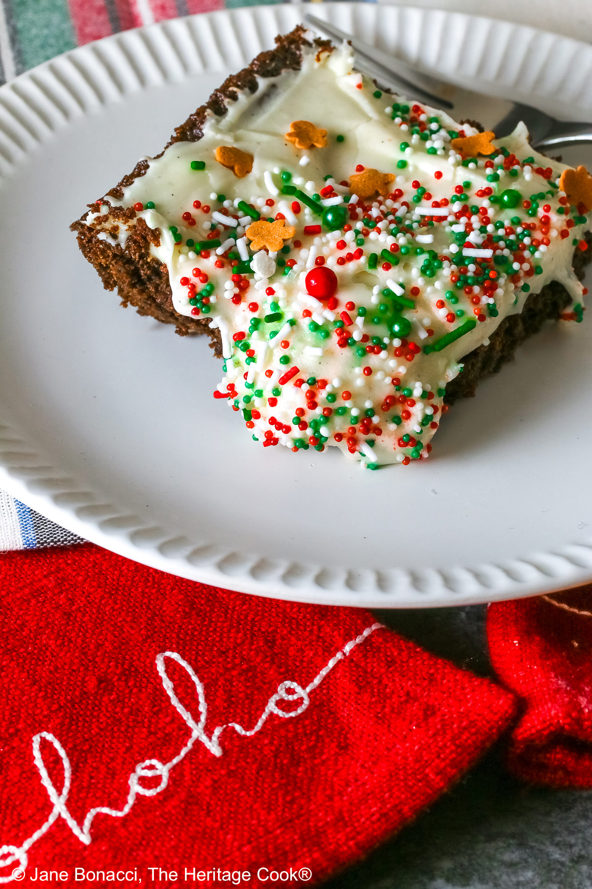 A slice of Gingerbread Brownies with Eggnog Frosting on a white plate with ridges at the edges and topped with green, red and white sprinkles with mini gingerbread men cookies, sitting on a festive holiday plaid patterned cloth and two red cloths with the phrases “ho ho ho” and “fa la la” stitched on them ©2025 Jane Bonacci, The Heritage Cook.