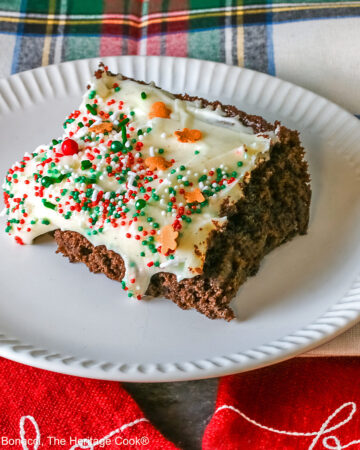 A slice of Gingerbread Brownies with Eggnog Frosting on a white plate with ridges at the edges and topped with green, red and white sprinkles with mini gingerbread men cookies, sitting on a festive holiday plaid patterned cloth and two red cloths with the phrases “ho ho ho” and “fa la la” stitched on them ©2025 Jane Bonacci, The Heritage Cook.