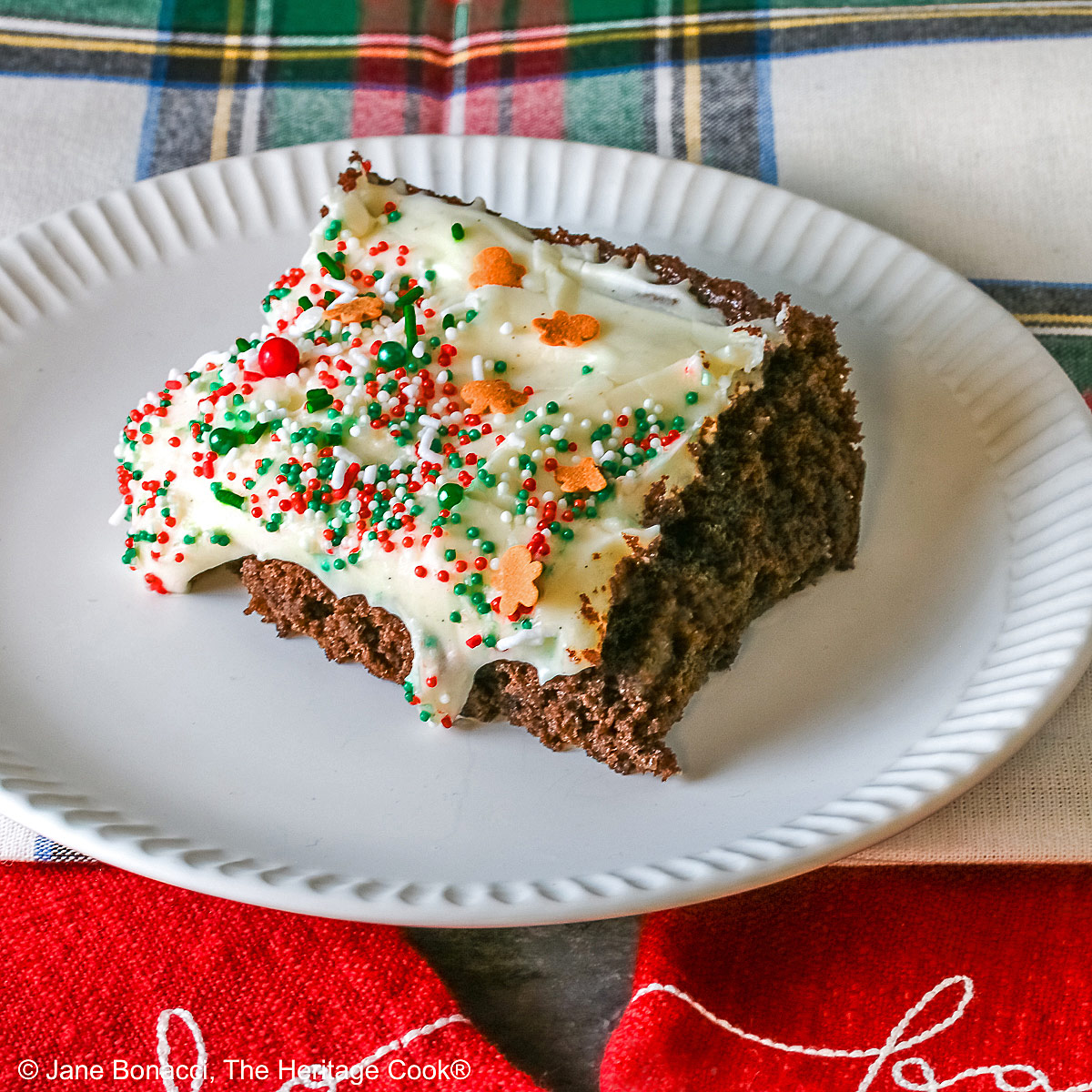 A slice of Gingerbread Brownies with Eggnog Frosting on a white plate with ridges at the edges and topped with green, red and white sprinkles with mini gingerbread men cookies, sitting on a festive holiday plaid patterned cloth and two red cloths with the phrases “ho ho ho” and “fa la la” stitched on them ©2025 Jane Bonacci, The Heritage Cook.