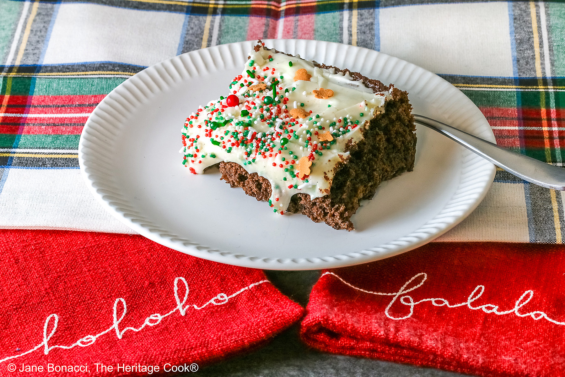 A slice of Gingerbread Brownies with Eggnog Frosting on a white plate with ridges at the edges and topped with green, red and white sprinkles with mini gingerbread men cookies, sitting on a festive holiday plaid patterned cloth and two red cloths with the phrases “ho ho ho” and “fa la la” stitched on them ©2025 Jane Bonacci, The Heritage Cook.