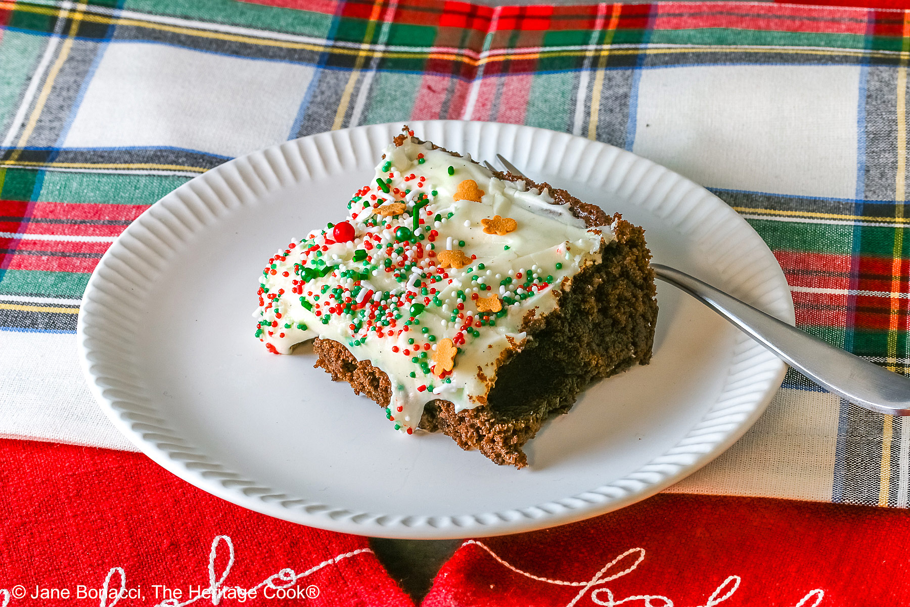 A slice of Gingerbread Brownies with Eggnog Frosting on a white plate with ridges at the edges and topped with green, red and white sprinkles with mini gingerbread men cookies, sitting on a festive holiday plaid patterned cloth and two red cloths with the phrases “ho ho ho” and “fa la la” stitched on them ©2025 Jane Bonacci, The Heritage Cook.