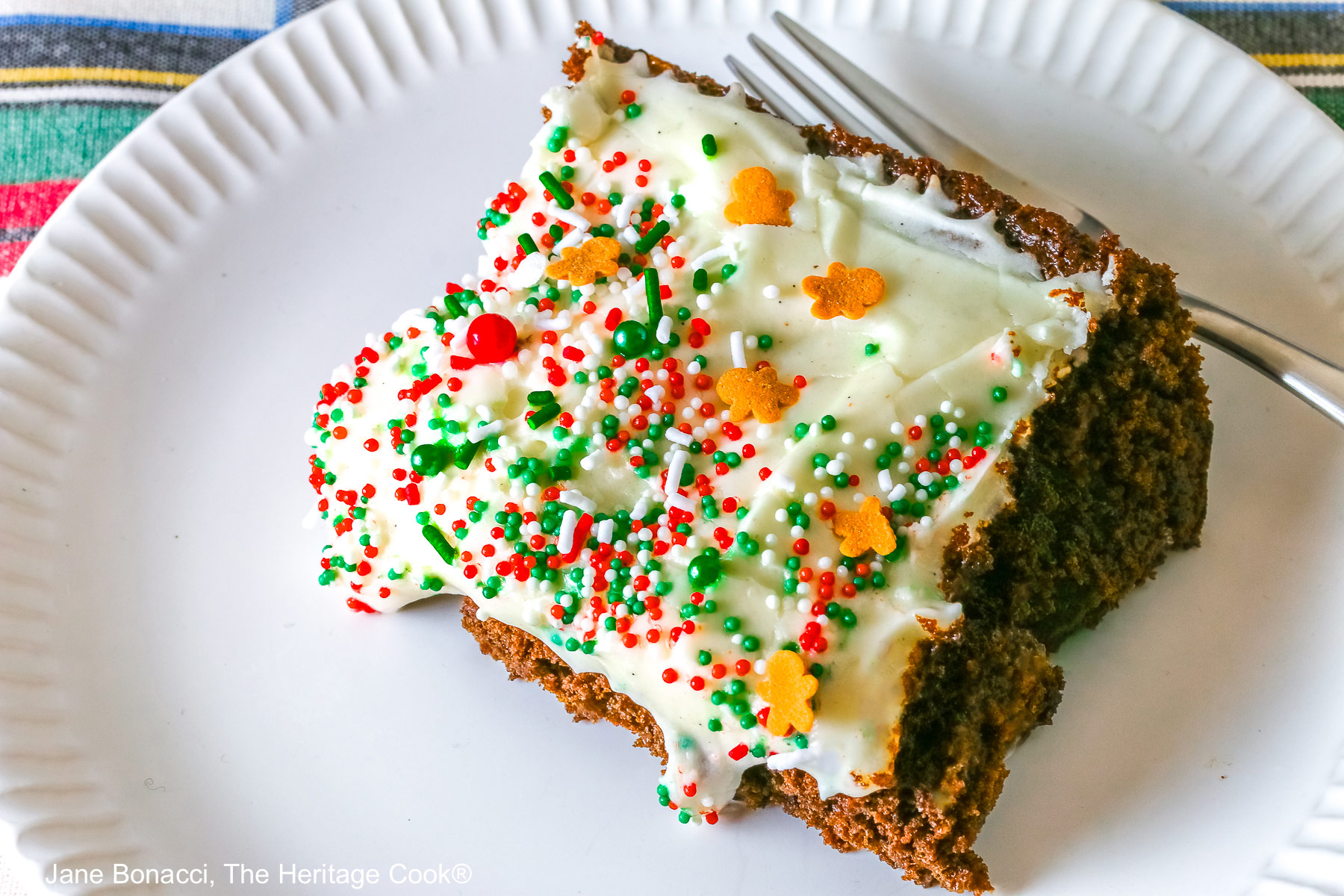 A slice of Gingerbread Brownies with Eggnog Frosting on a white plate with ridges at the edges and topped with green, red and white sprinkles with mini gingerbread men cookies, sitting on a festive holiday plaid patterned cloth and two red cloths with the phrases “ho ho ho” and “fa la la” stitched on them ©2025 Jane Bonacci, The Heritage Cook.