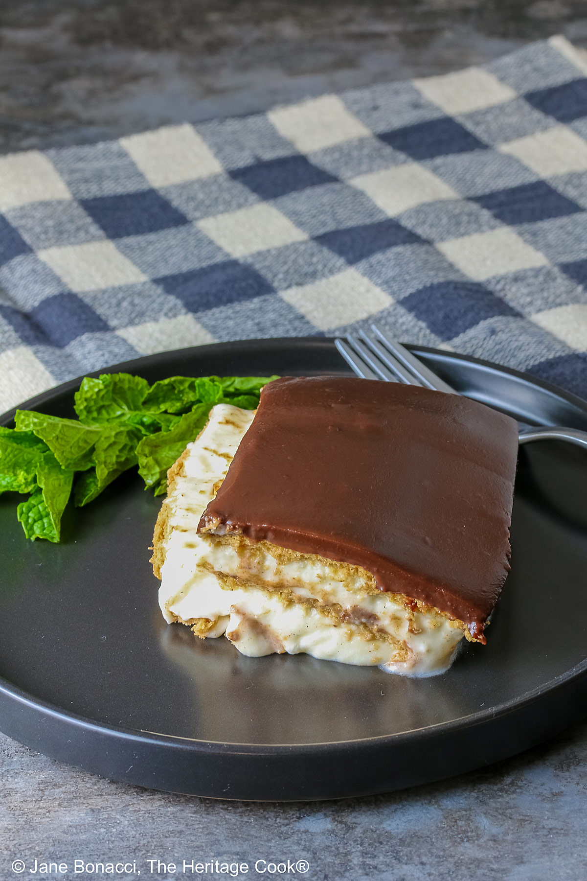 A square slice of the No Bake Chocolate Éclair Cake on a black plate with a sprig of mint and fork alongside, sitting on a muted blue and white checked cloth © 2025 Jane Bonacci, The Heritage Cook. 