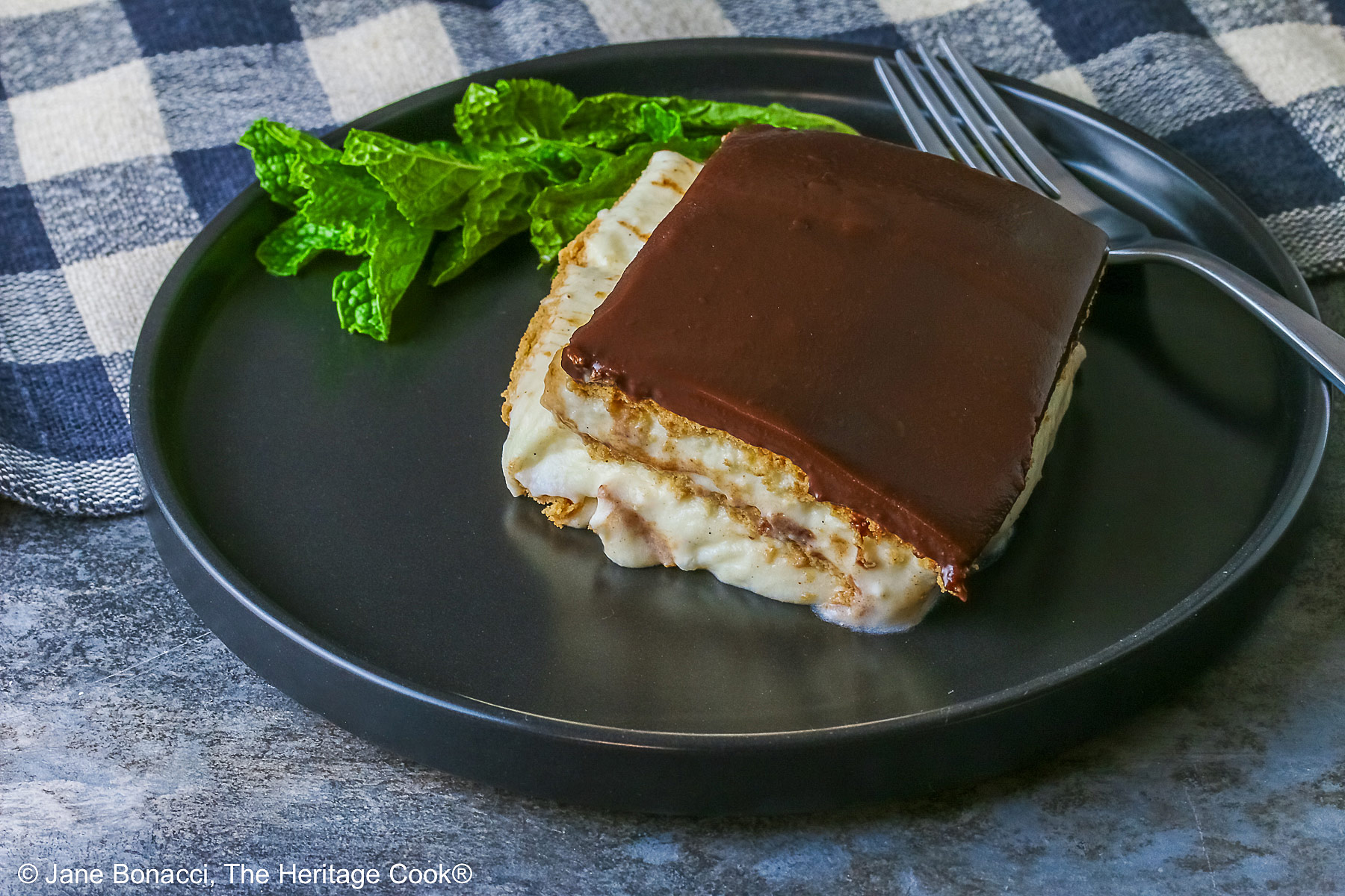 A square slice of the No Bake Chocolate Éclair Cake on a black plate with a sprig of mint and fork alongside, sitting on a muted blue and white checked cloth © 2025 Jane Bonacci, The Heritage Cook. 