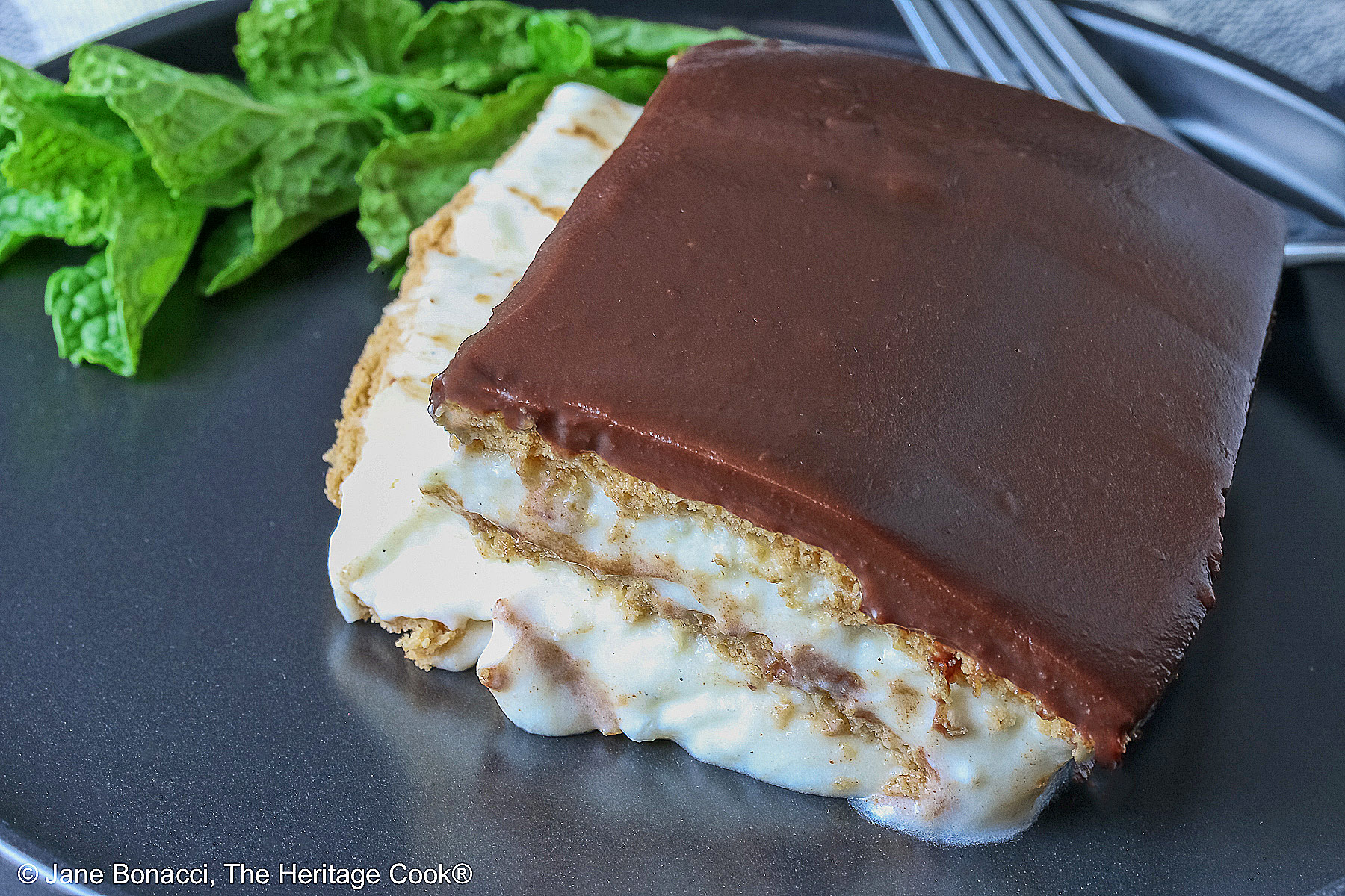 A square slice of the No Bake Chocolate Éclair Cake on a black plate with a sprig of mint and fork alongside, sitting on a muted blue and white checked cloth © 2025 Jane Bonacci, The Heritage Cook. 