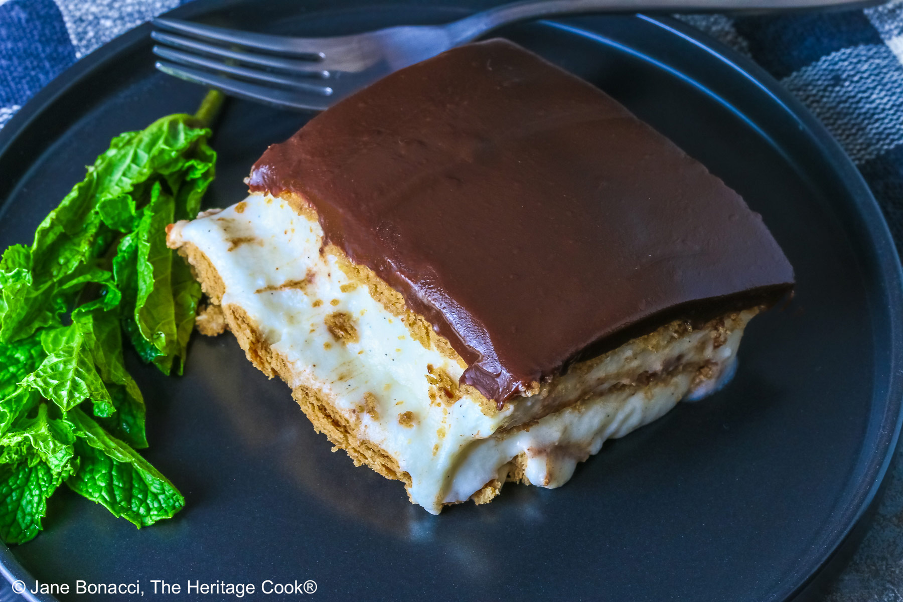 A square slice of the No Bake Chocolate Éclair Cake on a black plate with a sprig of mint and fork alongside, sitting on a muted blue and white checked cloth © 2025 Jane Bonacci, The Heritage Cook. 