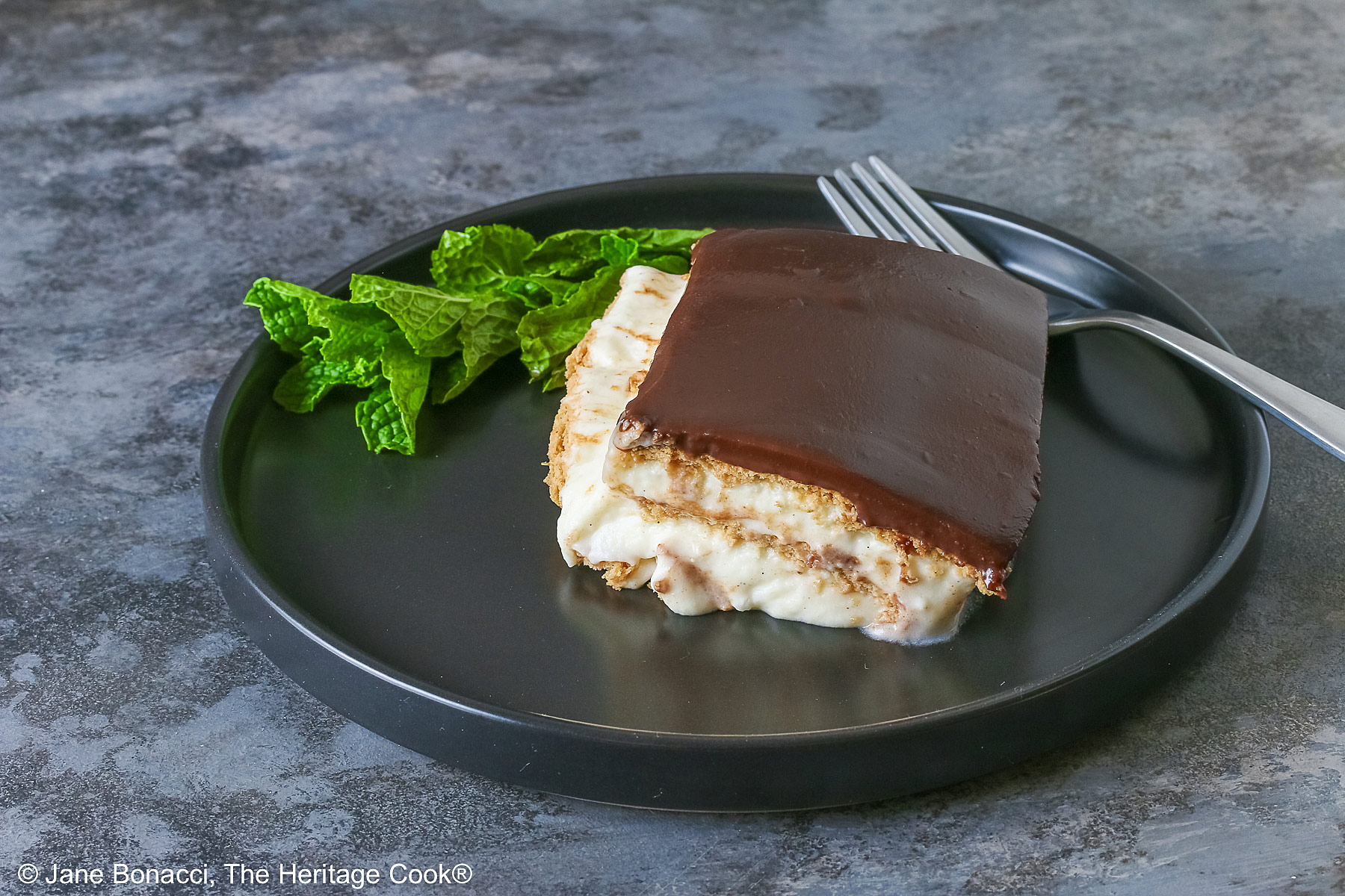 A square slice of the No Bake Chocolate Éclair Cake on a black plate with a sprig of mint and fork alongside, sitting on a muted blue and white checked cloth © 2025 Jane Bonacci, The Heritage Cook. 