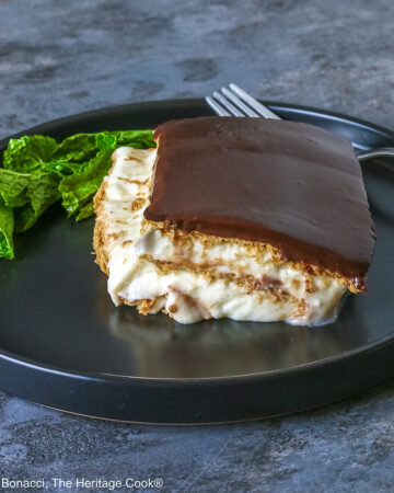 A square slice of the No Bake Chocolate Éclair Cake on a black plate with a sprig of mint and fork alongside, sitting on a muted blue and white checked cloth © 2025 Jane Bonacci, The Heritage Cook.