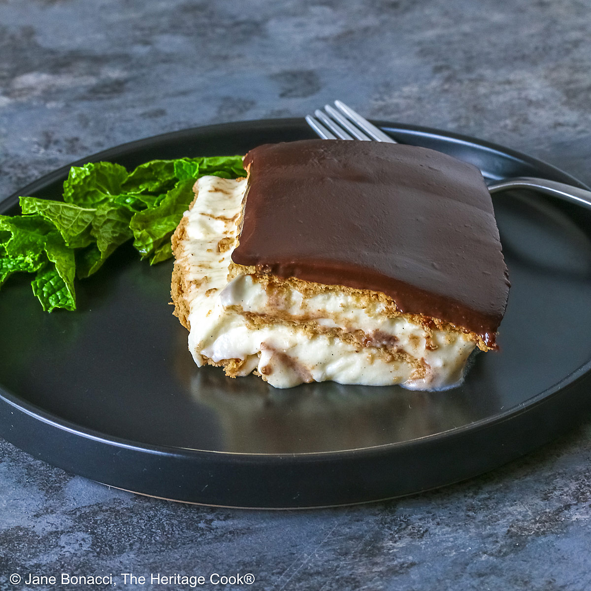 A square slice of the No Bake Chocolate Éclair Cake on a black plate with a sprig of mint and fork alongside, sitting on a muted blue and white checked cloth © 2025 Jane Bonacci, The Heritage Cook. 