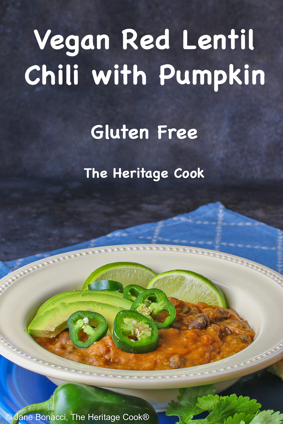 Pale yellow bowl of Vegan Red Lentil Chili with Pumpkin sitting on a blue plate surrounded by jalapeno peppers, cilantro, lime, and cornbread, with a blue cloth beneath © 2025 Jane Bonacci, The Heritage Cook